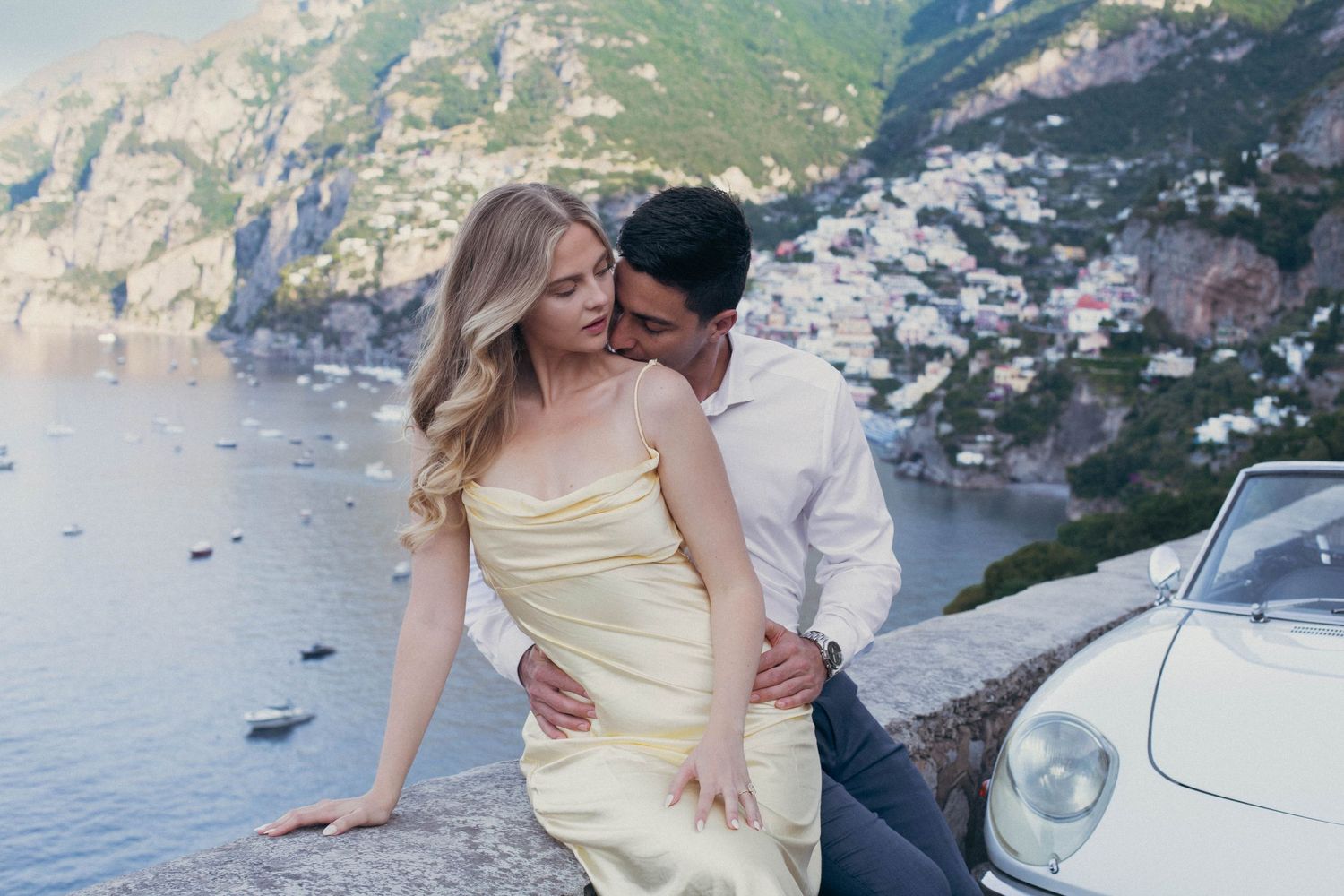 Romantic couple embracing by vintage car with Positano coastline and turquoise sea behind them.