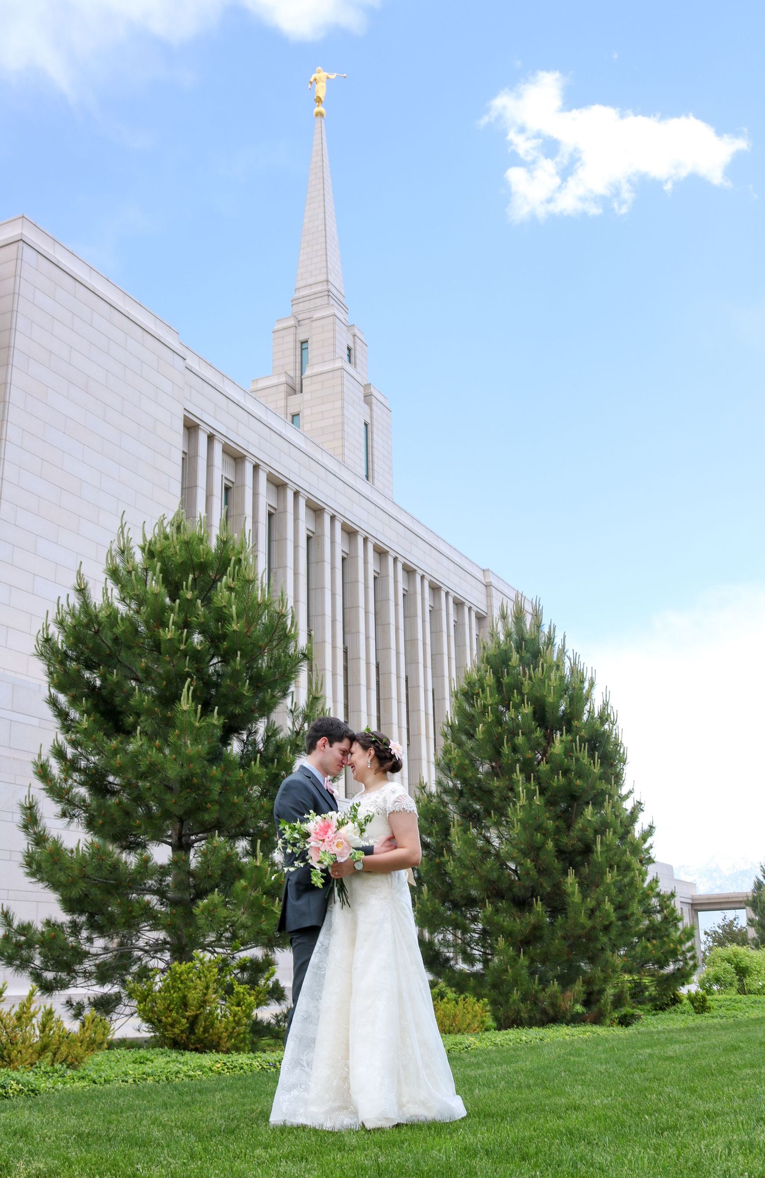 Couple in wedding attire posing on grassy hill with white temple and blue sky in background.