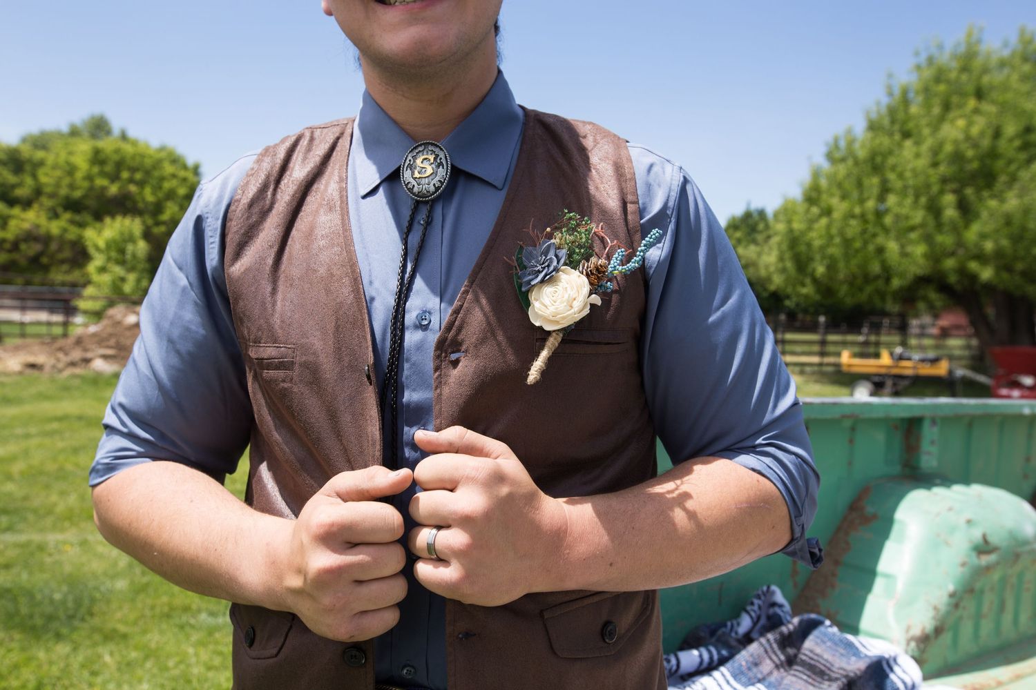 Close-up of groom wearing brown leather vest with bolo tie and white boutonniere on sunny wedding day.