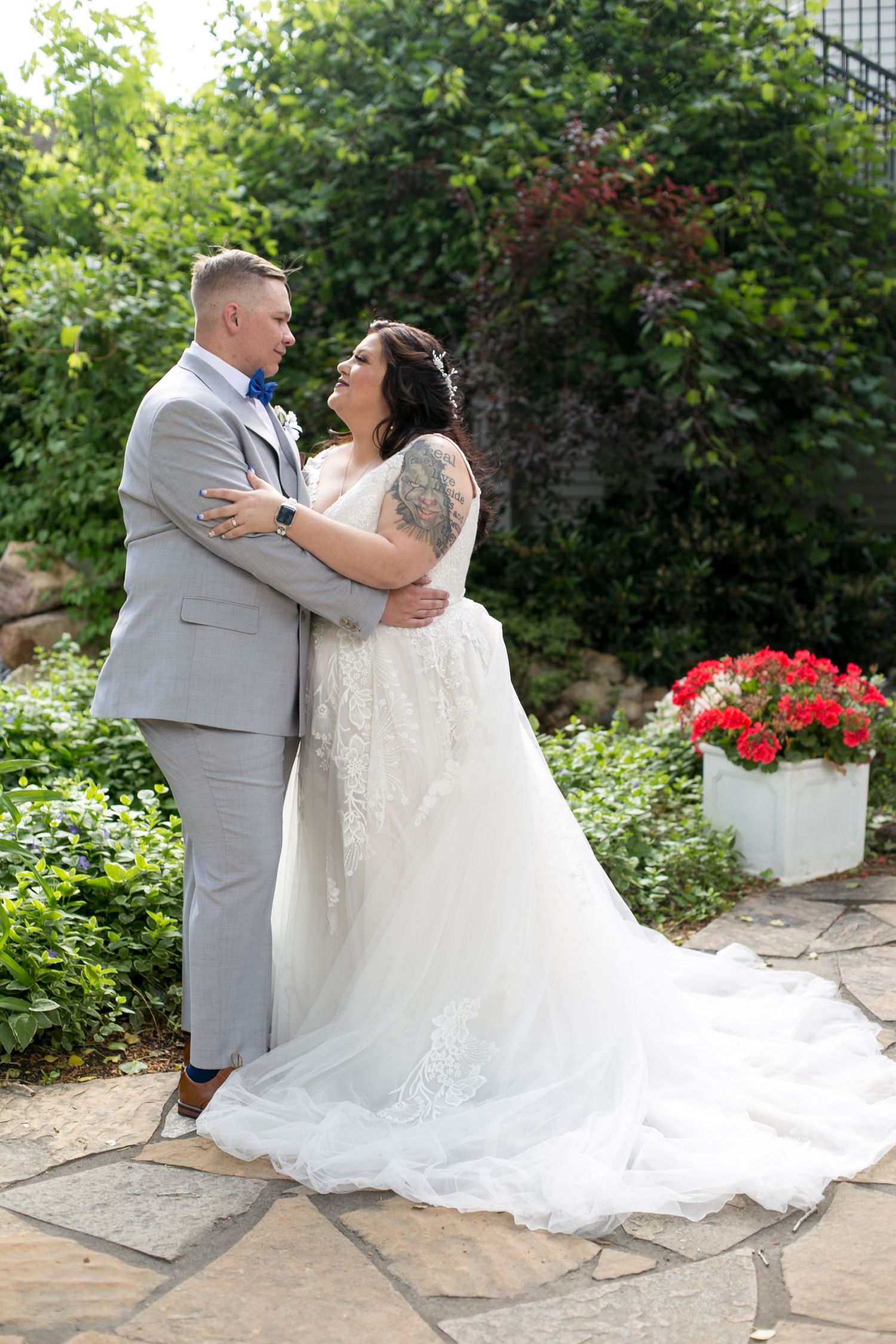 A bride and groom embrace outdoors on a stone patio surrounded by lush greenery and red flowers on their wedding day.