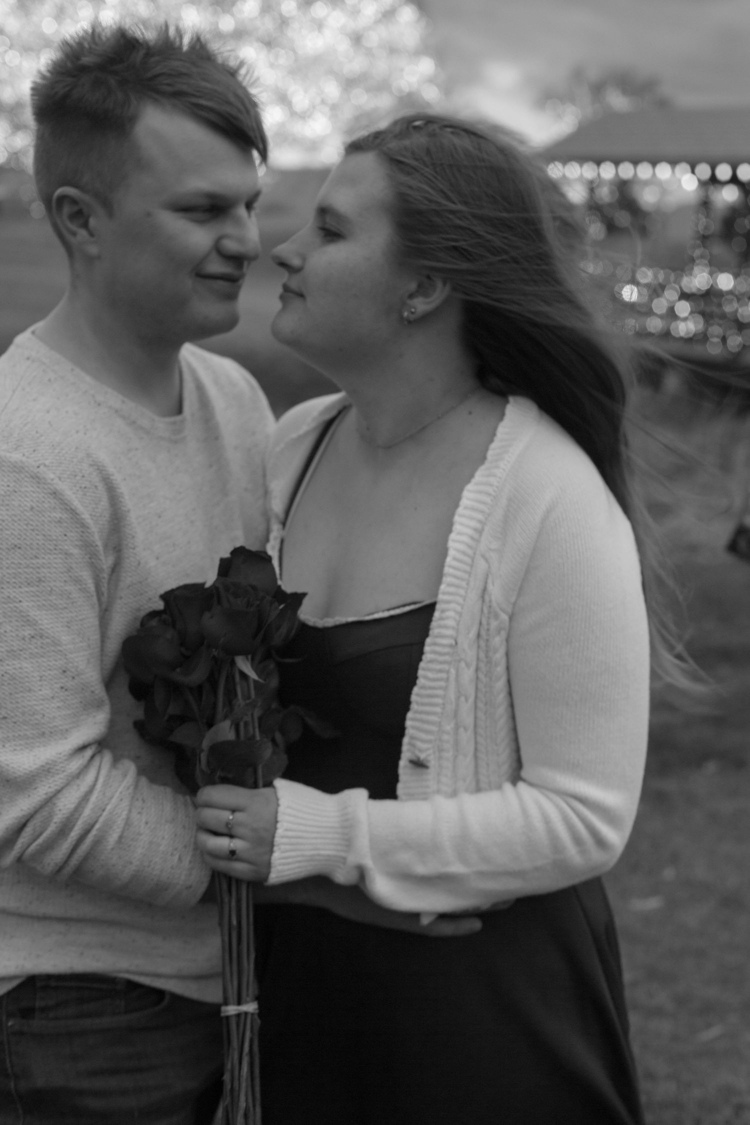 Black and white photo of a young couple sharing a romantic moment, man holding dark roses outdoors with bokeh lights.