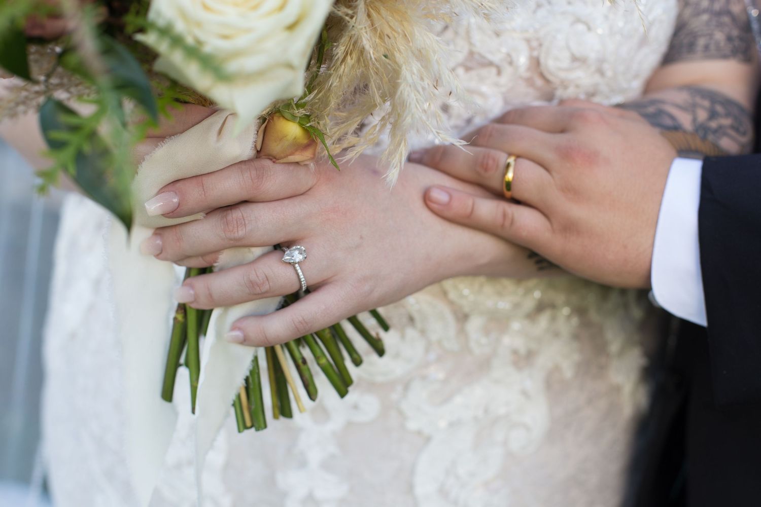 Close-up of wedding rings and hands holding a white rose bouquet with green stems.