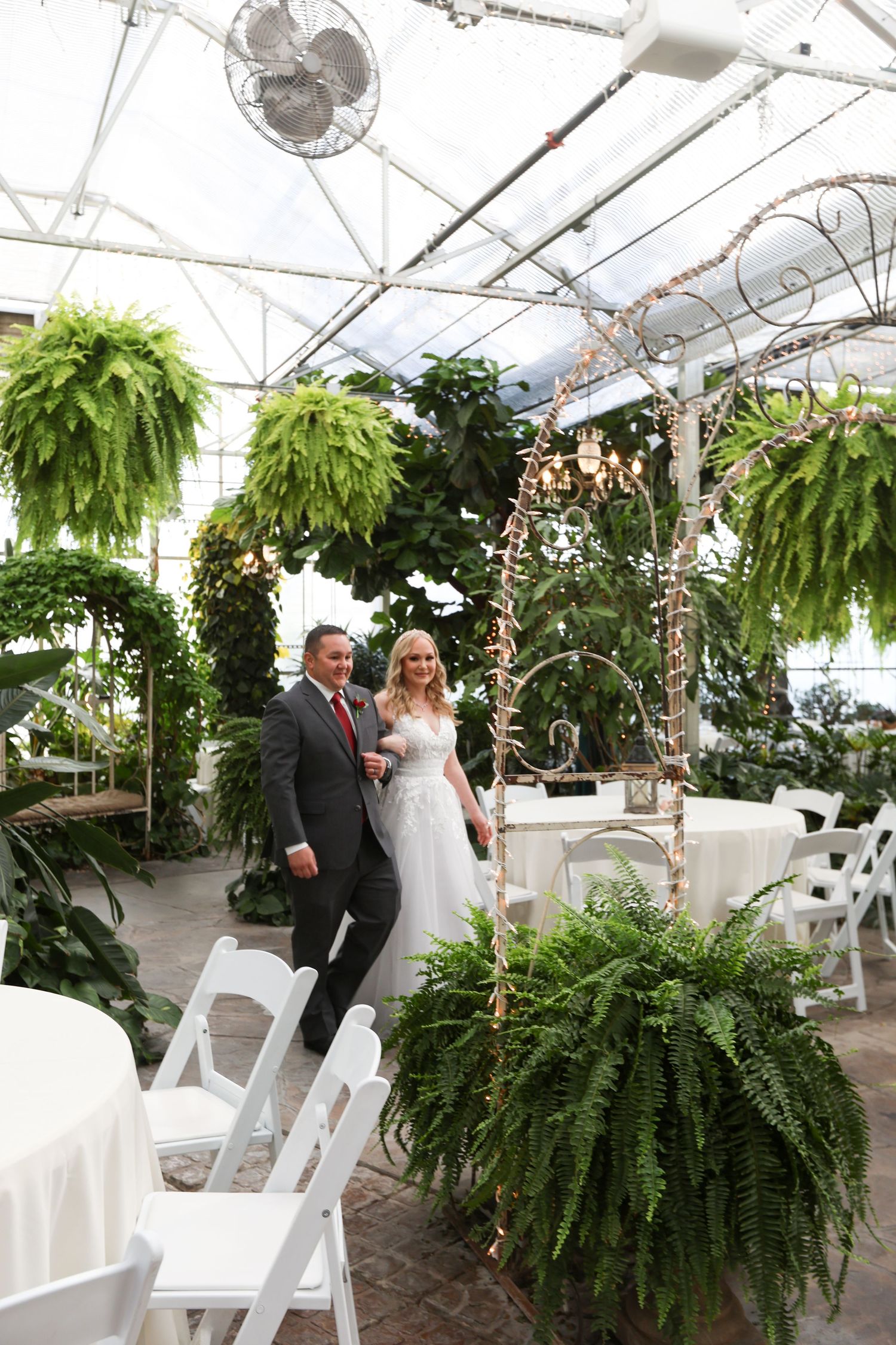 Wedding ceremony setup in a greenhouse with hanging ferns and white folding chairs.