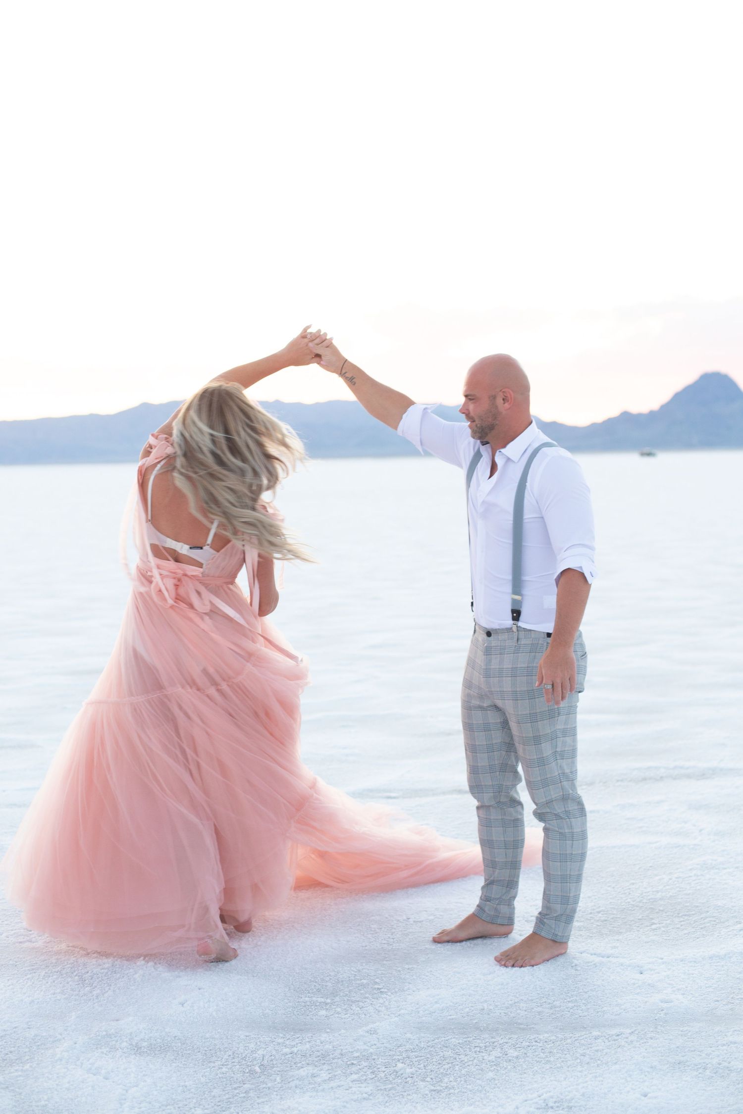 Couple performs romantic dance moves on salt flats with mountain backdrop at sunset.