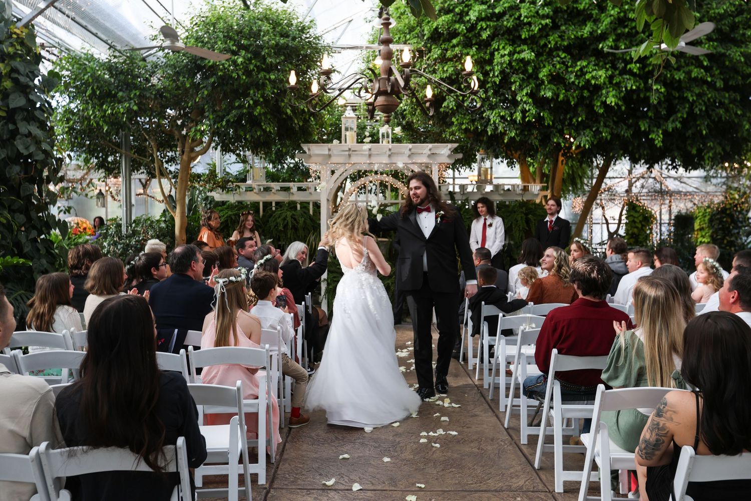 An outdoor wedding ceremony setup with white chairs and greenery in a glass-covered venue.