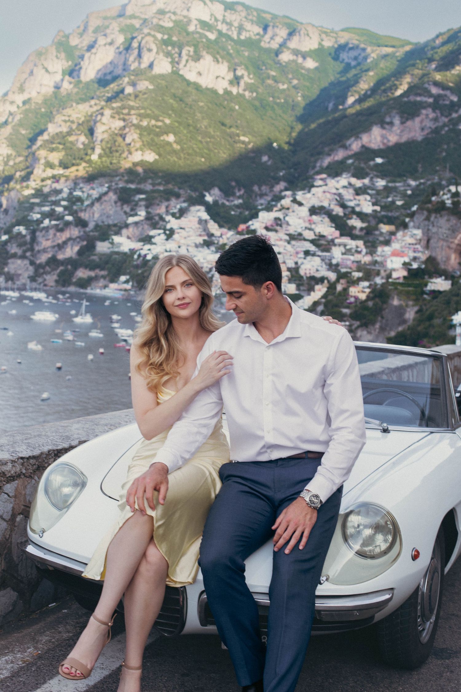 Couple posing by a vintage white convertible with Positano's colorful cliffside village and bay in the background.