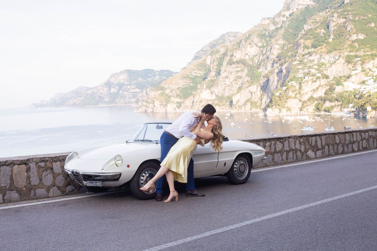 Couple sharing a romantic dip kiss beside white vintage convertible on the scenic Amalfi Coast highway.