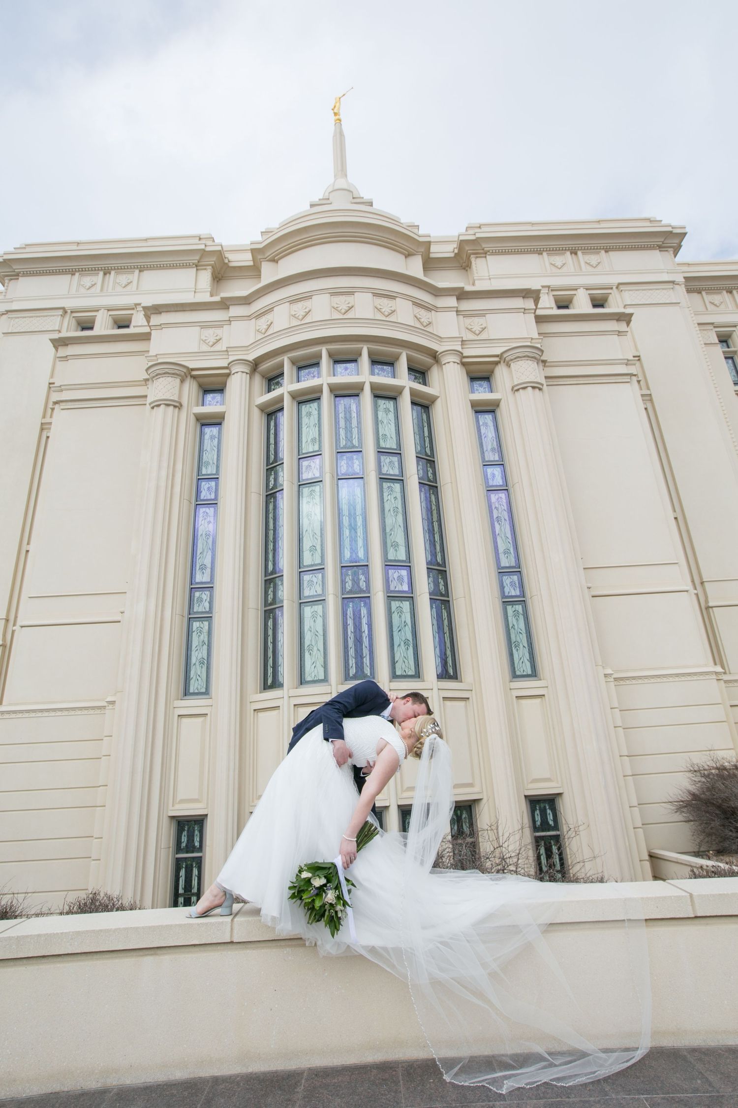 Groom dips bride in a romantic kiss outside a beautiful white LDS temple with tall stained glass windows.