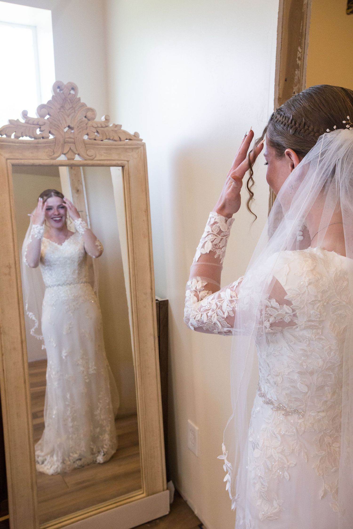 Bride in lace wedding gown adjusts veil while smiling at her reflection in an ornate gold floor mirror.