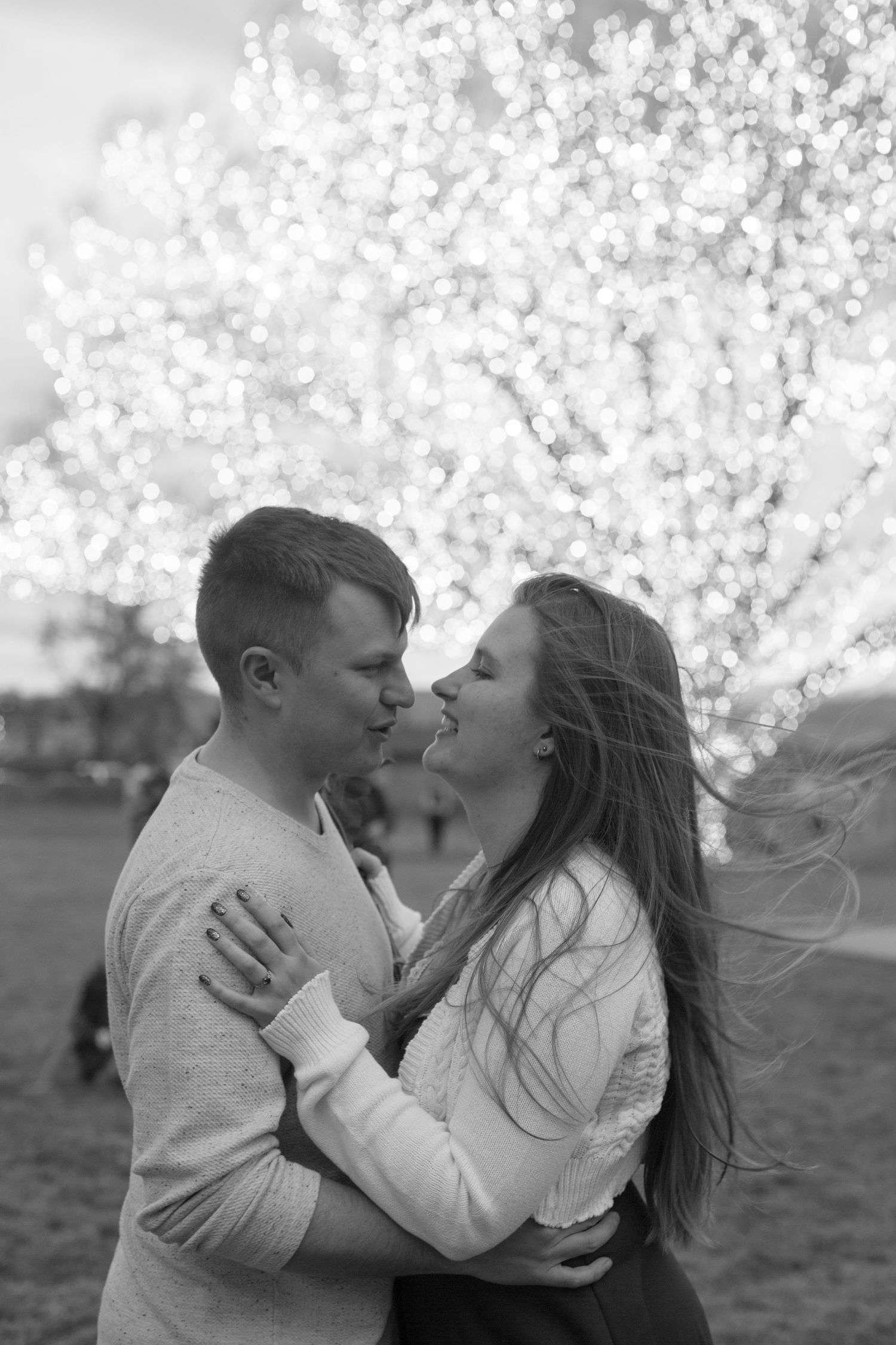 Black and white photo of a romantic couple embracing and about to kiss in front of a bokeh lit tree.