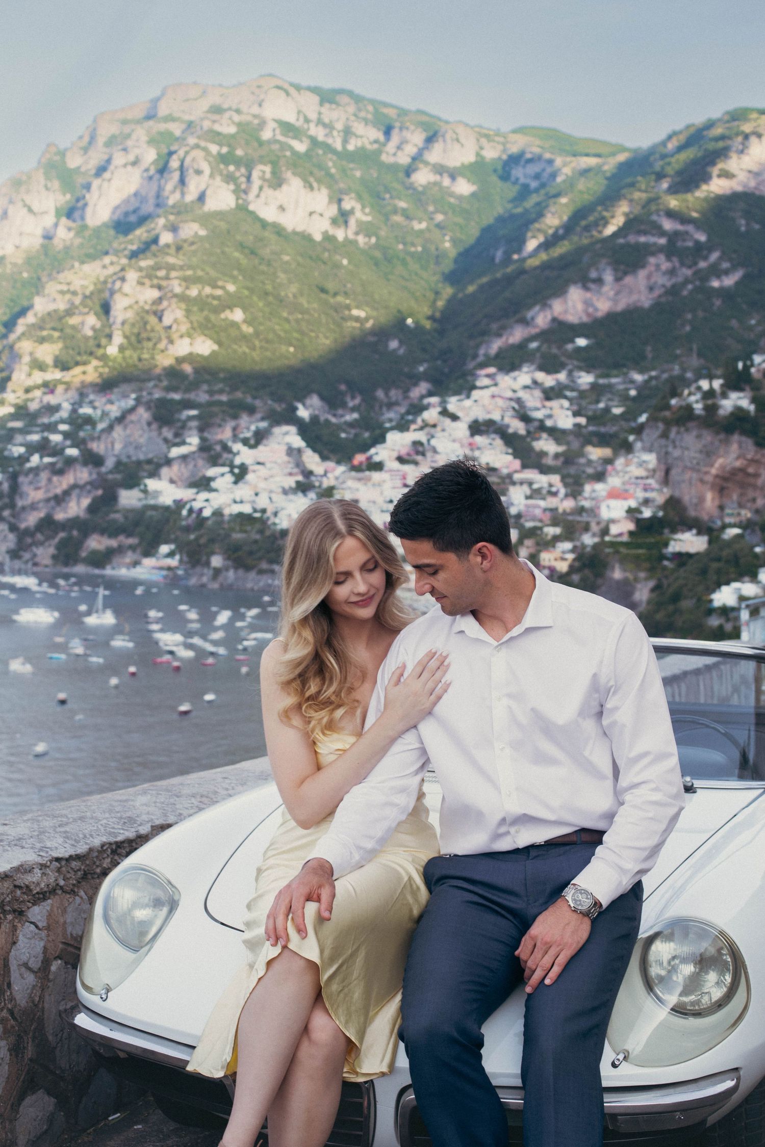 Romantic couple sitting on vintage white car with Positano cliffside village and Mediterranean sea backdrop.