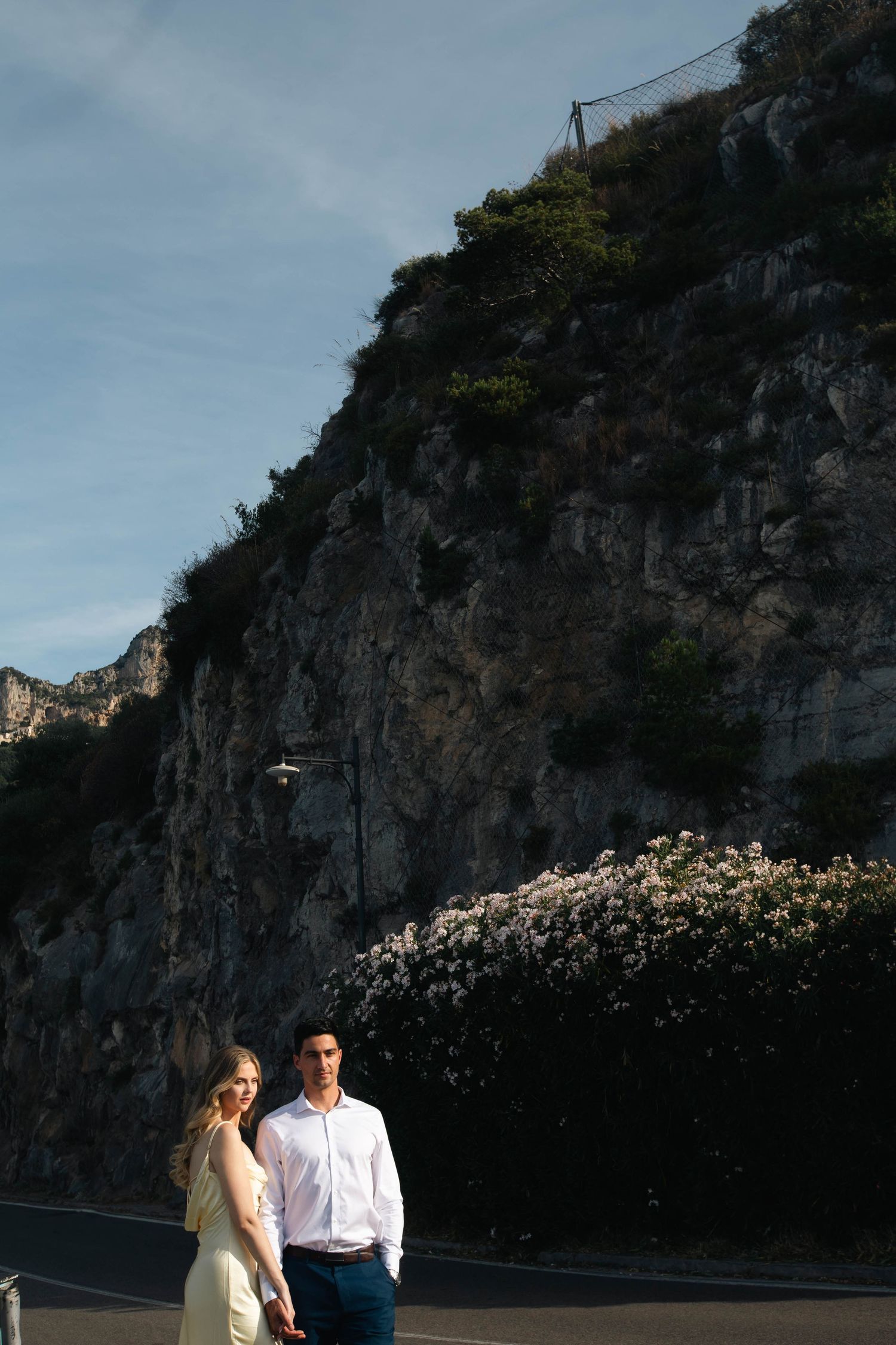 Couple holding hands on a sunlit road beneath dramatic rocky cliffs with blooming white flowers nearby.