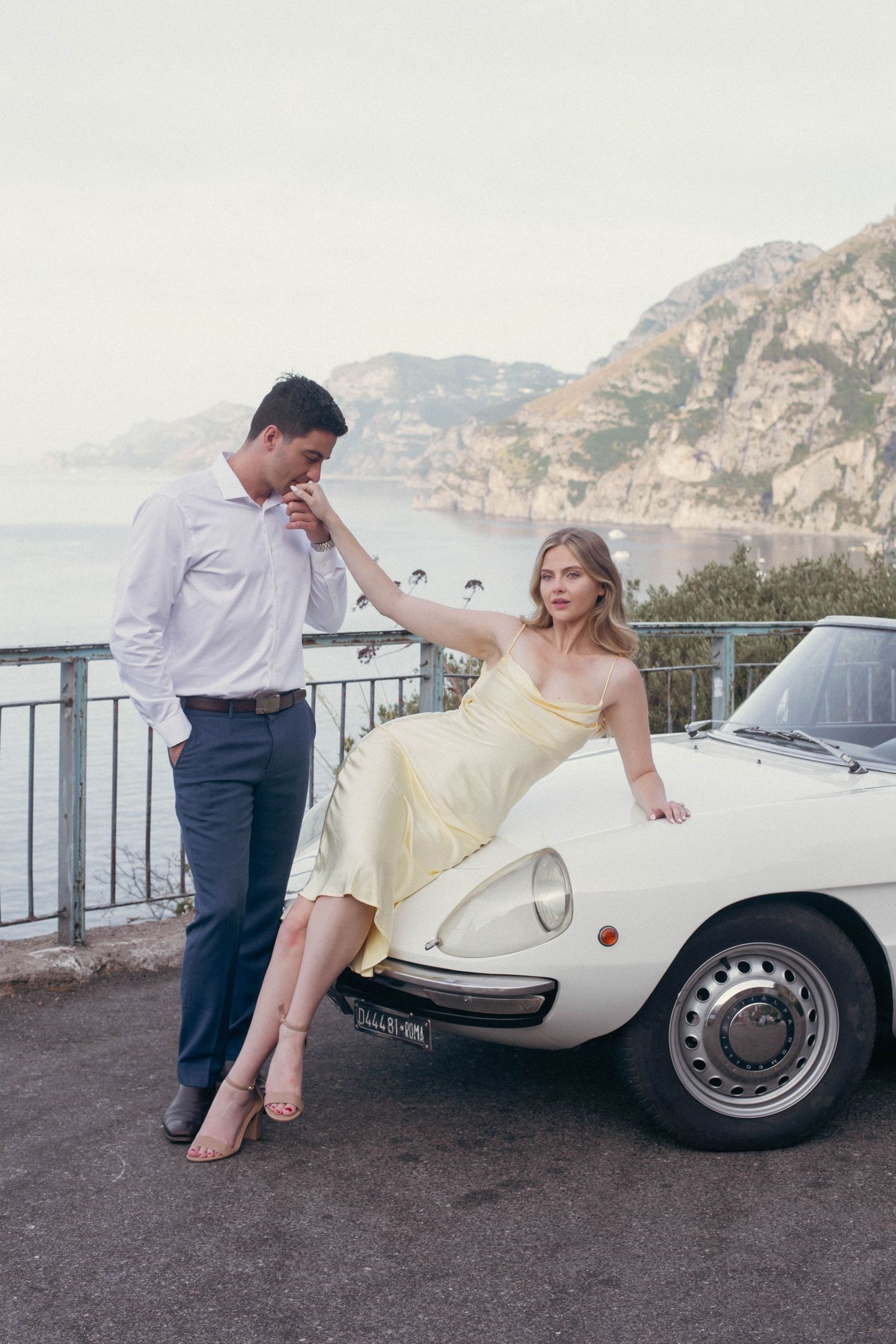 Man kissing womans hand as she sits on white classic Alfa Romeo Spider on the Amalfi Coast.