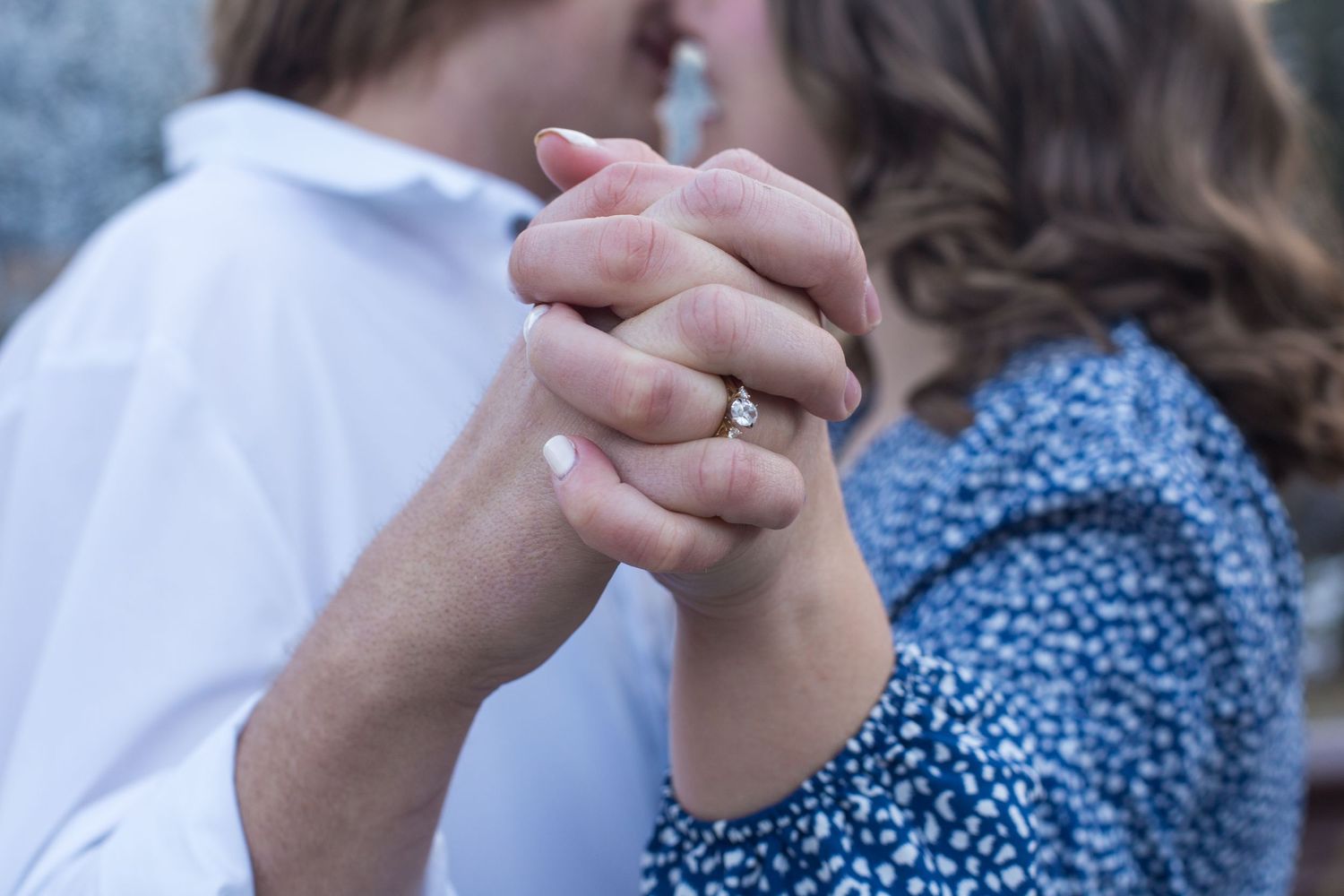 Close-up of clasped hands showing engagement ring against blue patterned dress.