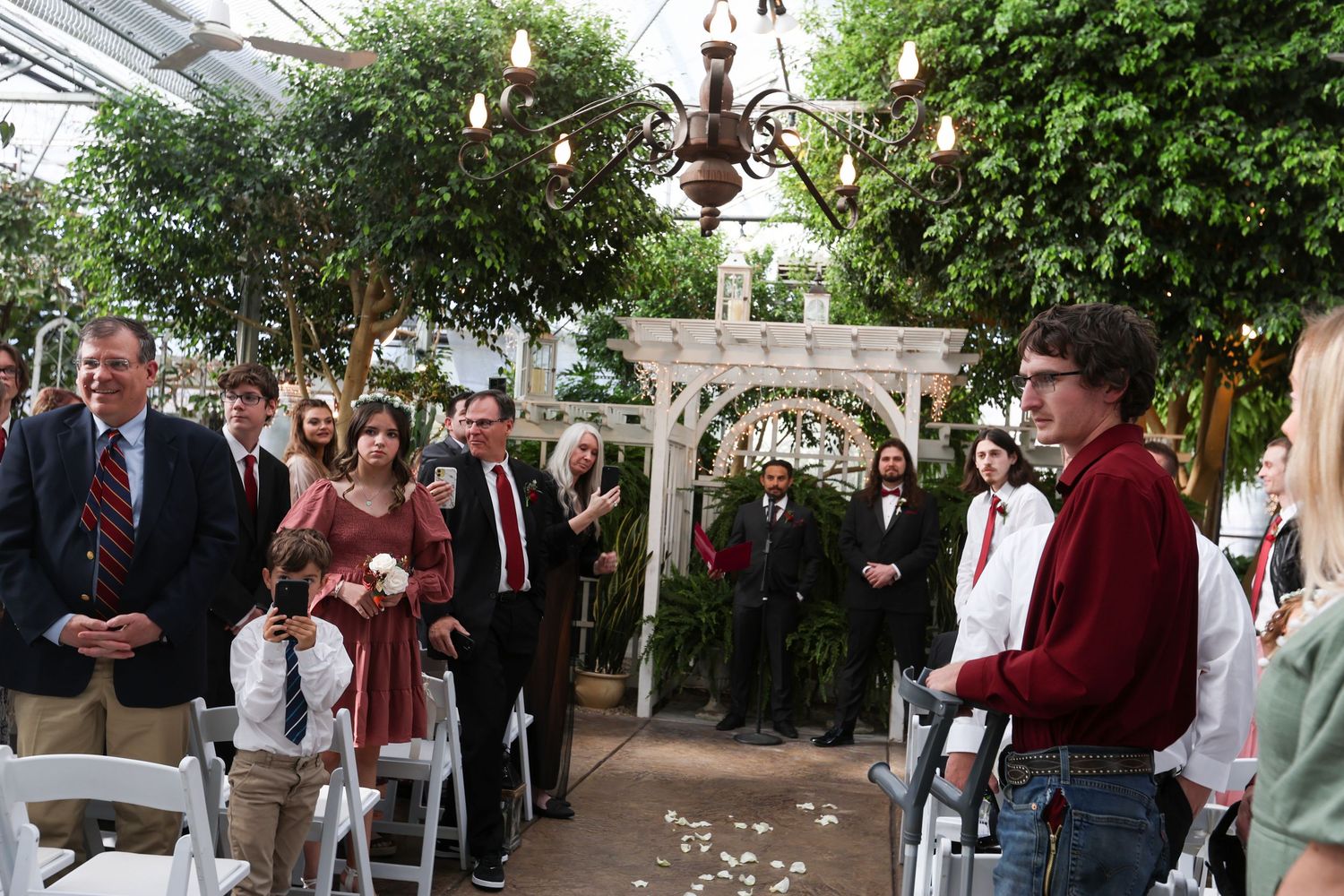 Wedding ceremony in progress inside a greenhouse with guests and musicians.