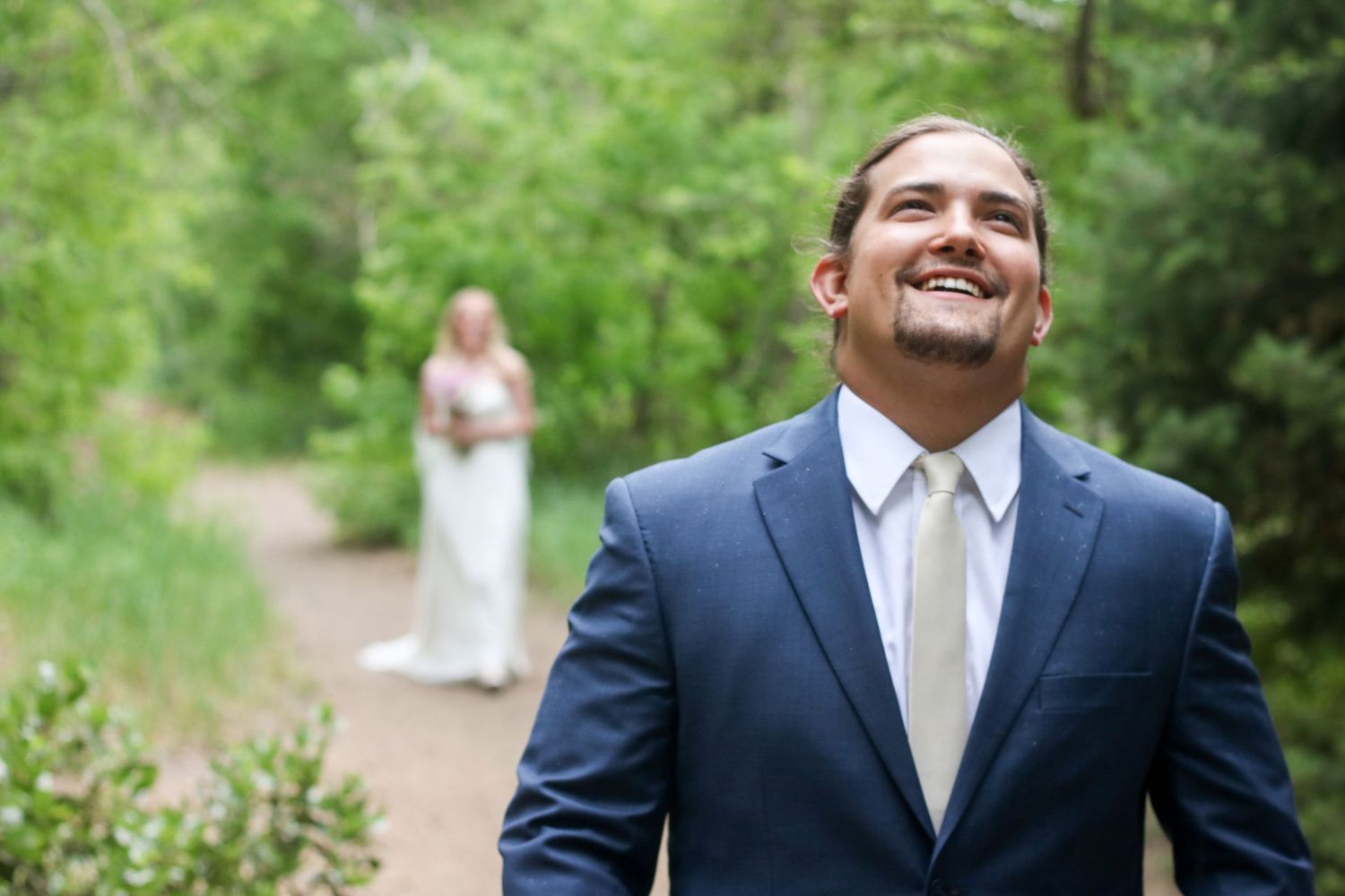 A groom in a navy suit smiles joyfully on a forest path during a wedding ceremony.