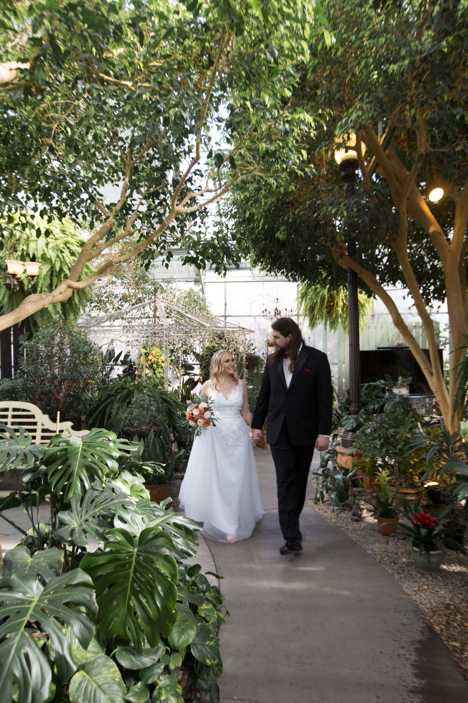 A couple walks through a lush garden pathway with tropical plants and hanging lights.