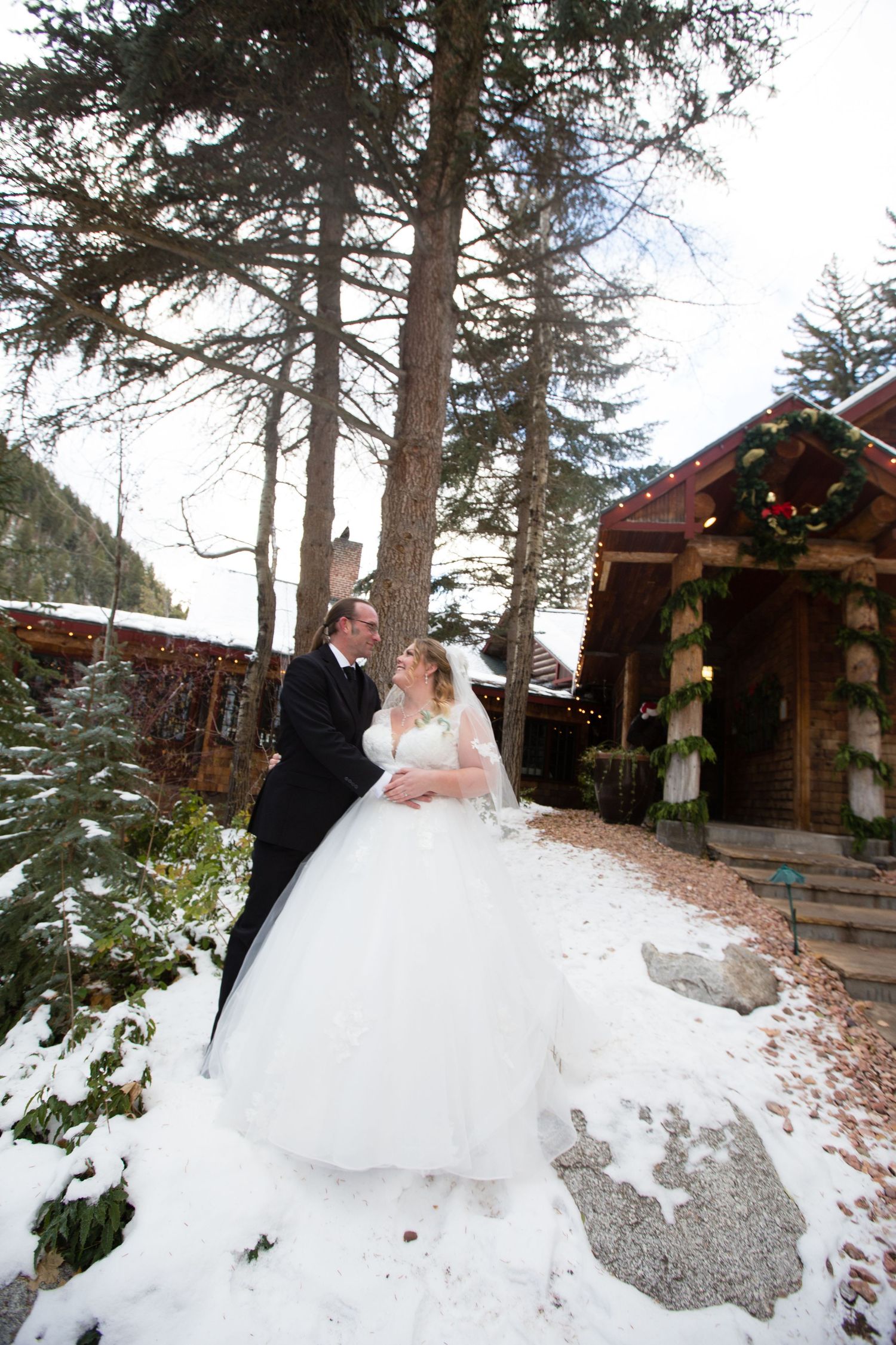 A romantic winter wedding portrait with rustic cabins and decorated evergreens in the background.