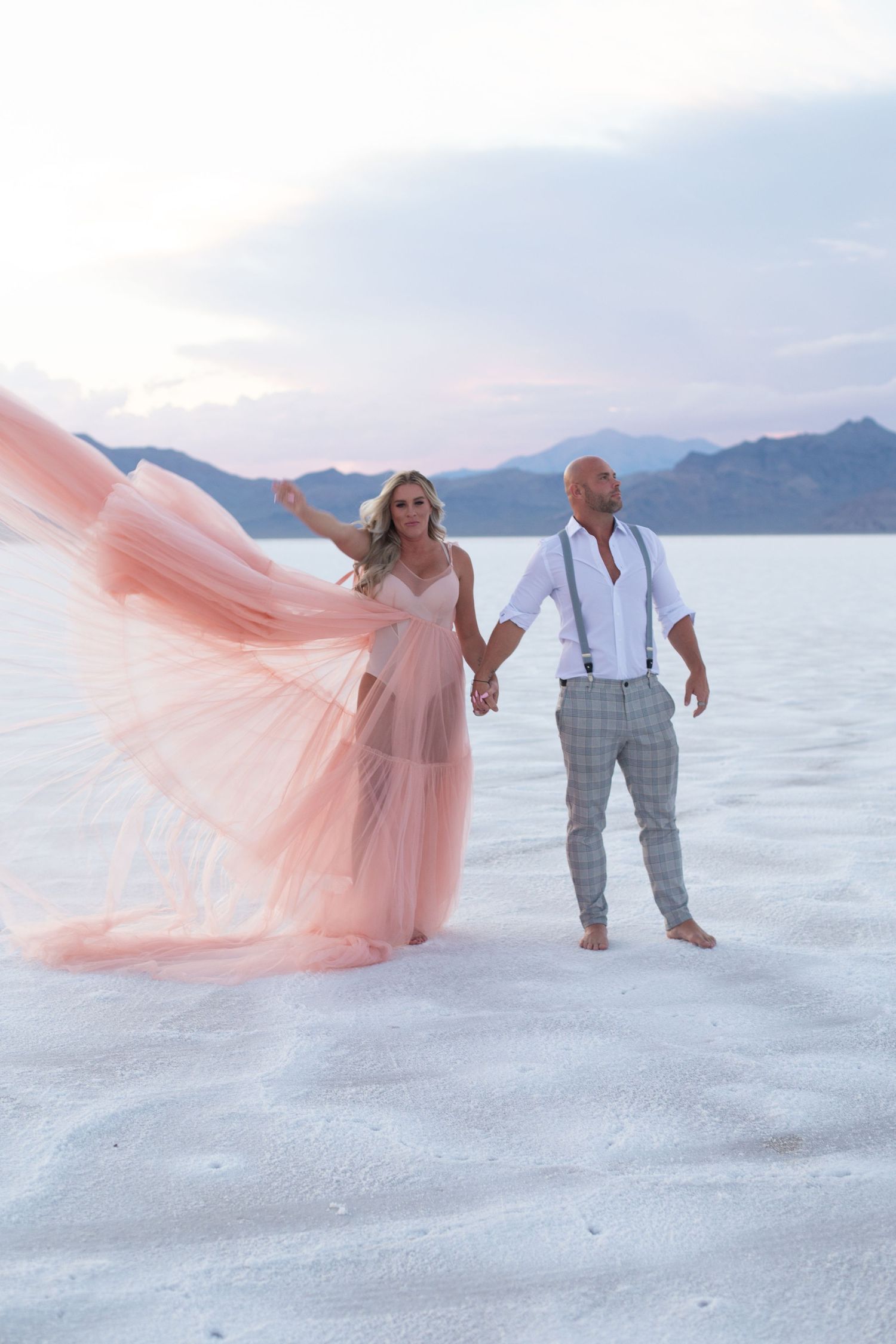 Couple walks hand in hand on salt flats, pink dress flowing in the wind.