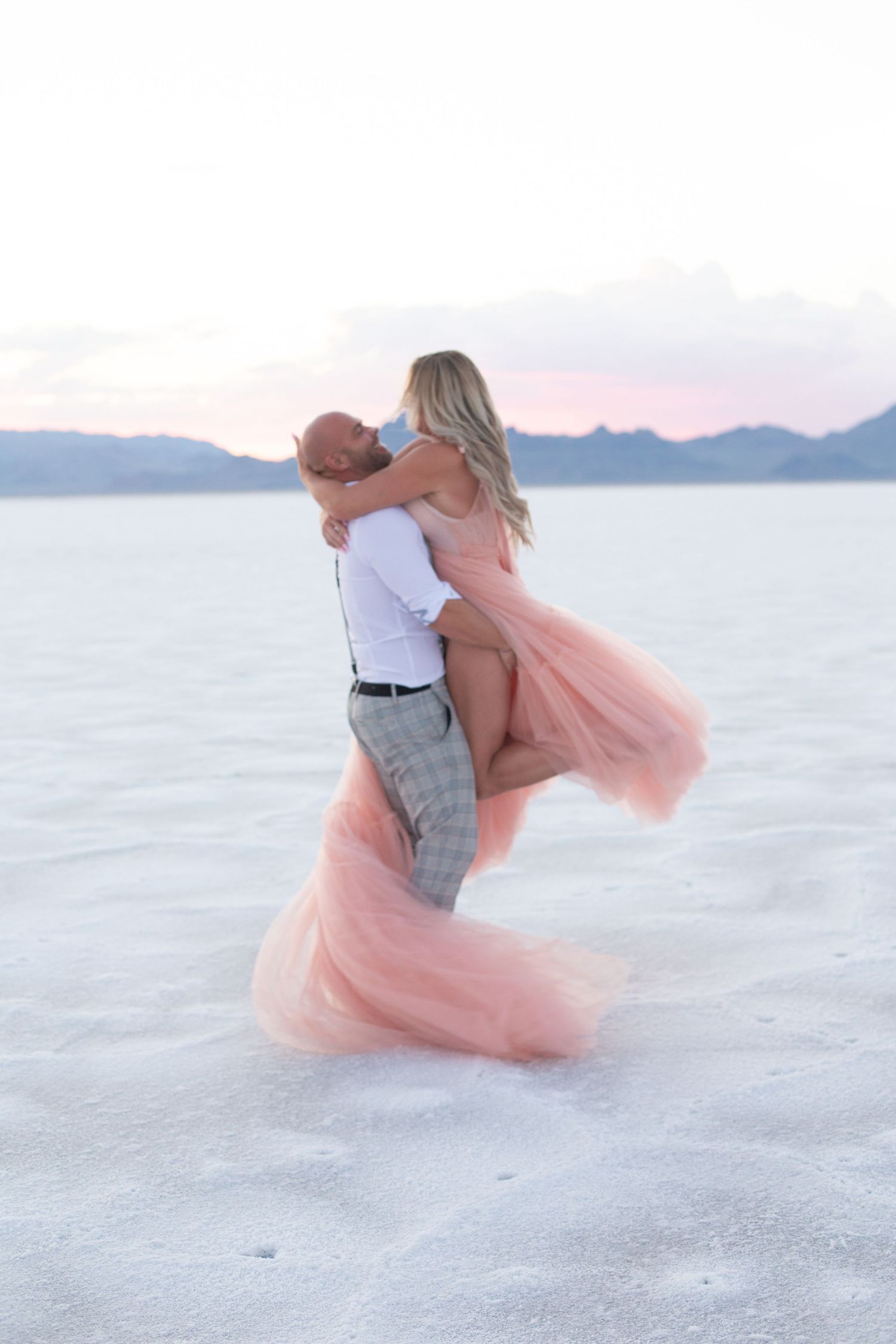 Playful lift pose on white salt flats with flowing pink dress against mountain backdrop.