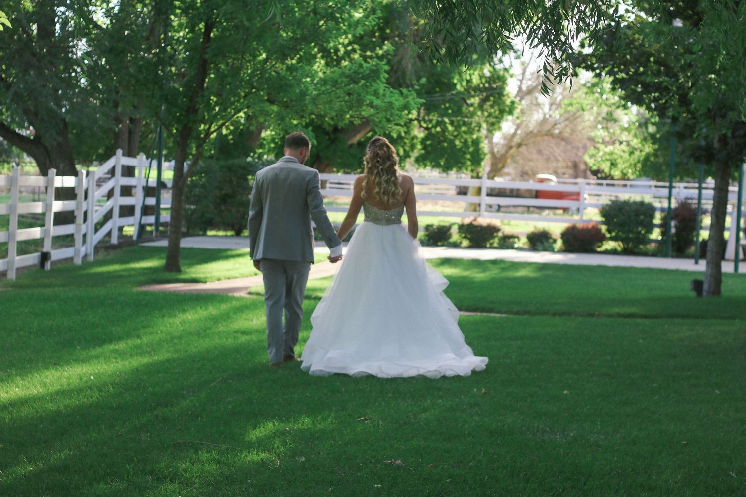 Couple walking along white fence on manicured lawn in wedding attire.