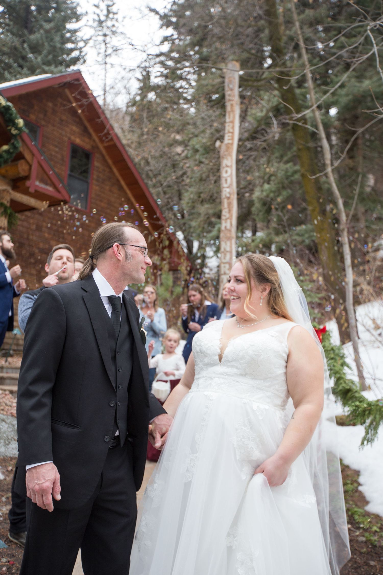 Bride and groom hold hands smiling at each other after winter wedding ceremony outside rustic log cabin with snow.