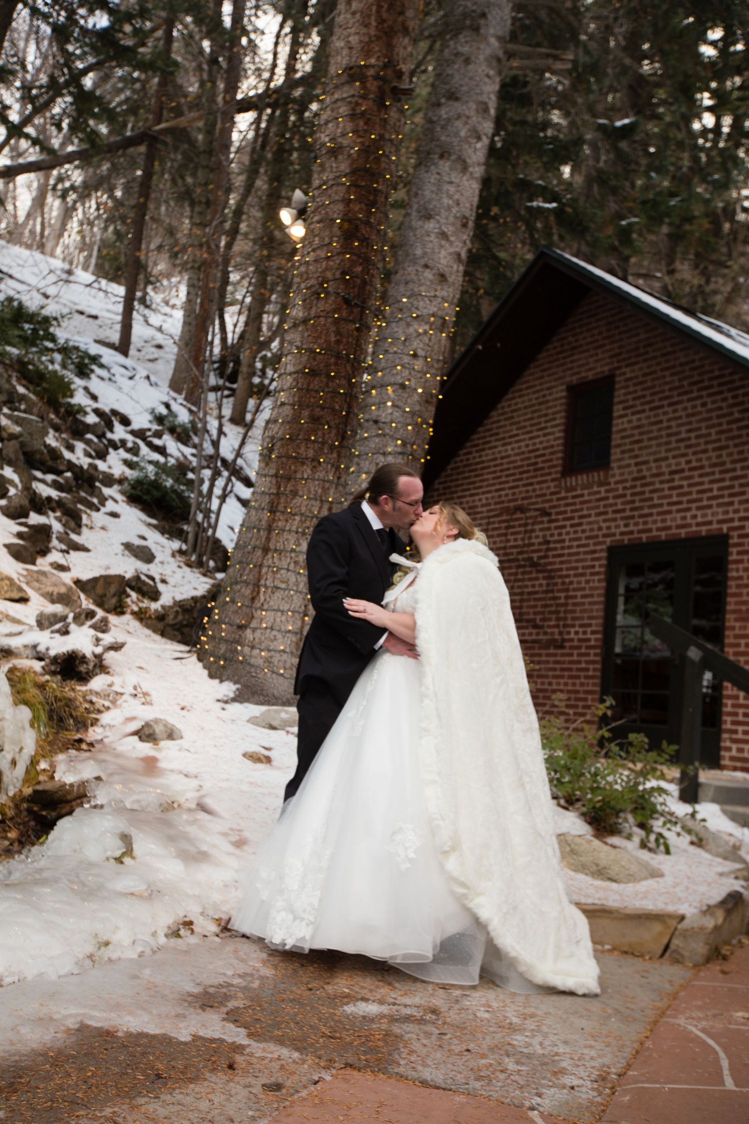 A romantic winter wedding photoshoot with a couple embracing near a brick building surrounded by snow and trees.