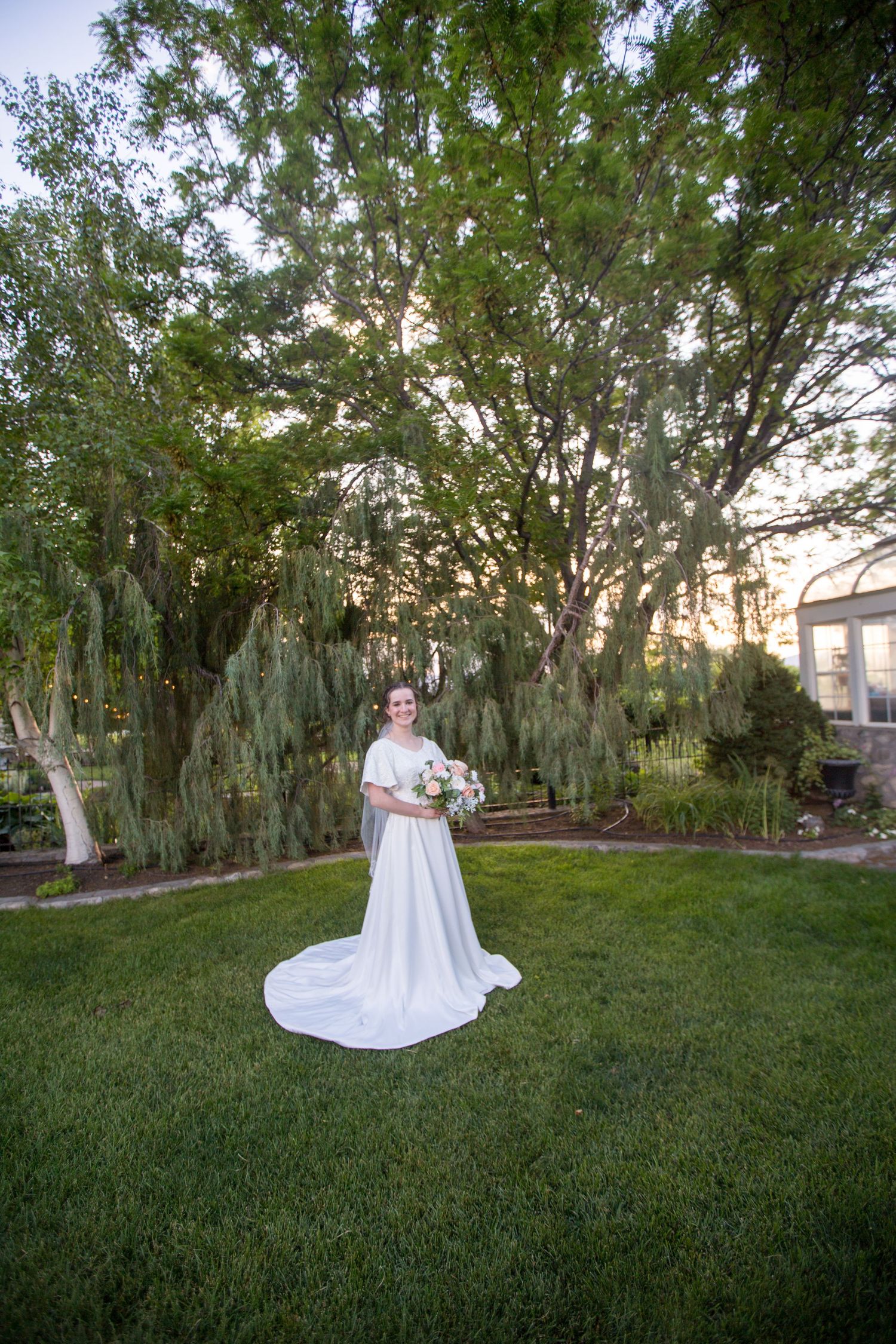 A white wedding dress flows across a green lawn surrounded by tall trees at sunset.