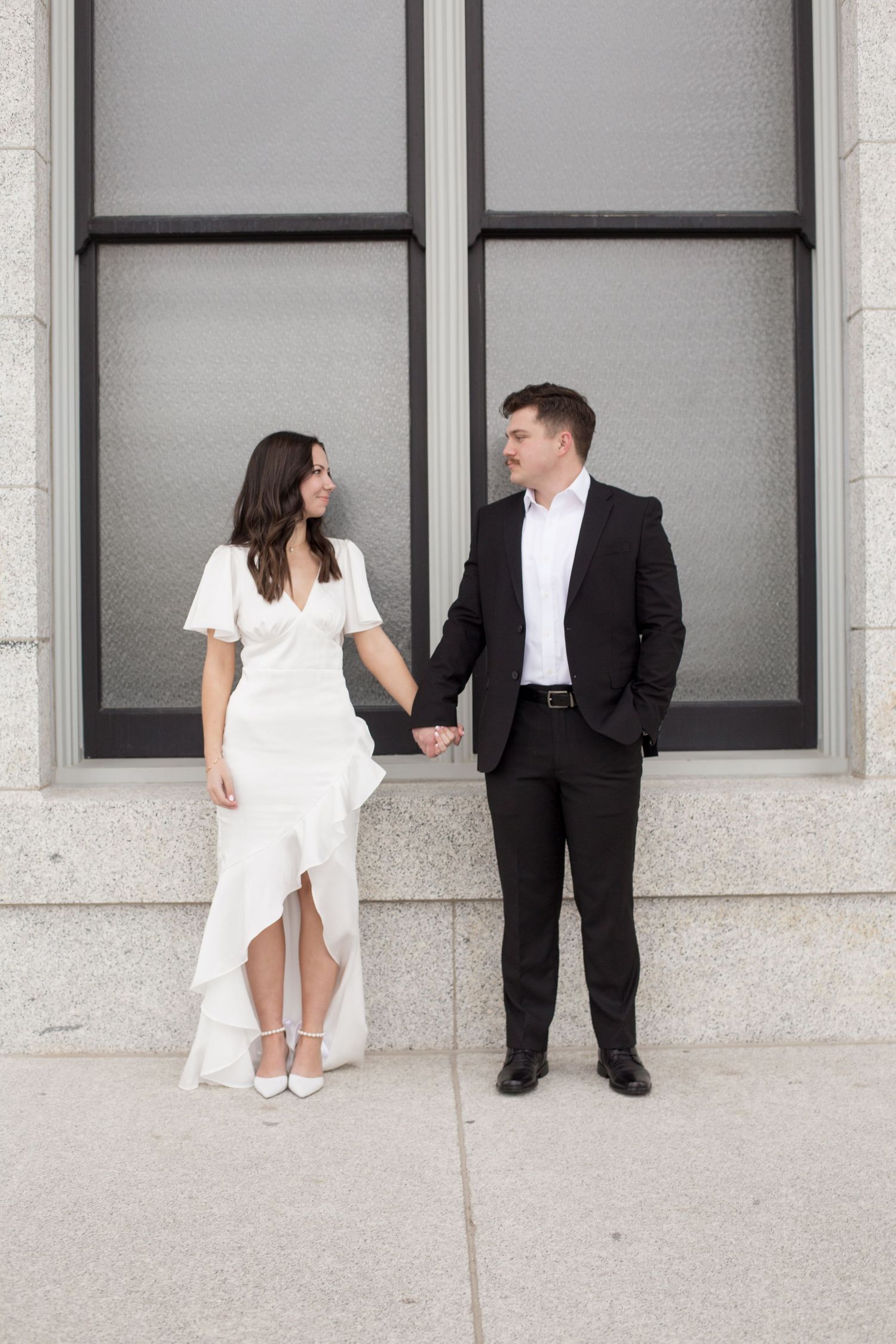 Couple in formal attire poses playfully against a modern building facade.