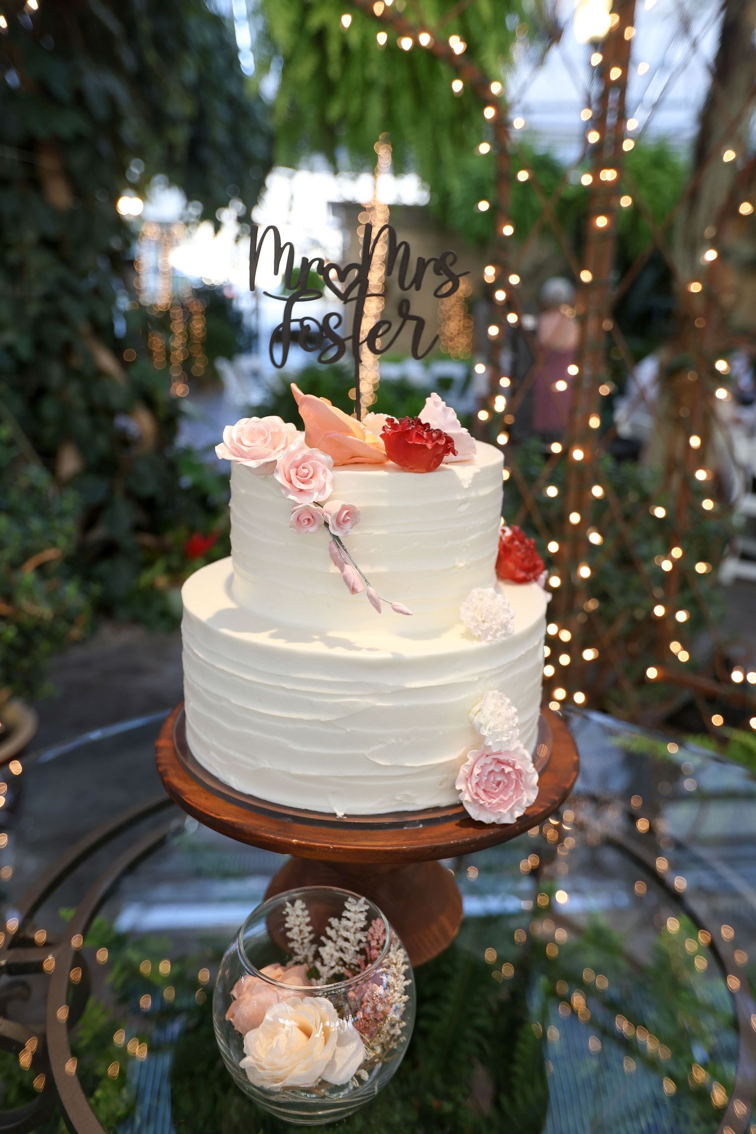 White wedding cake decorated with pink roses displayed with twinkle lights in background.