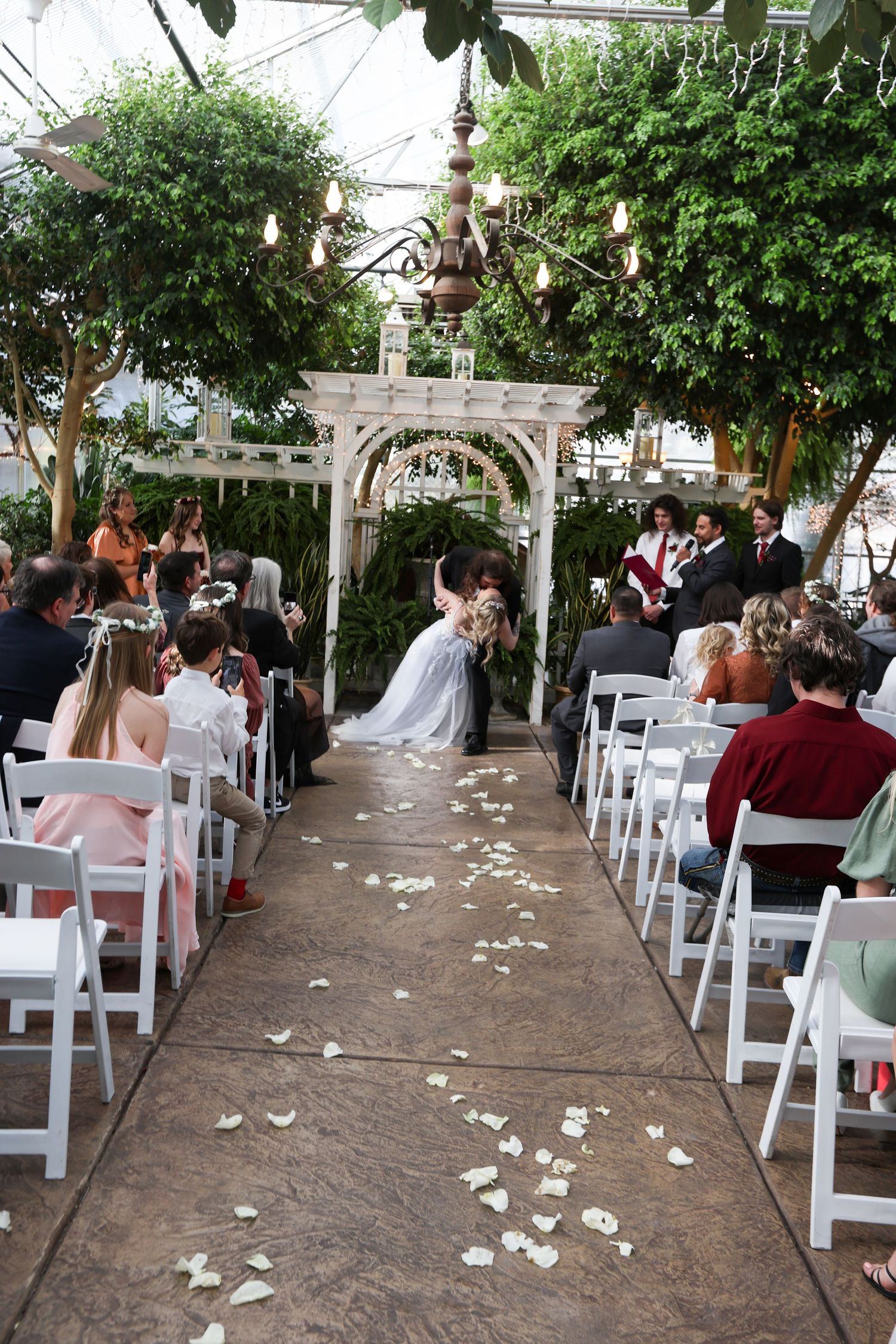 An outdoor wedding ceremony setup with white chairs and flower petals along a tree-lined path.