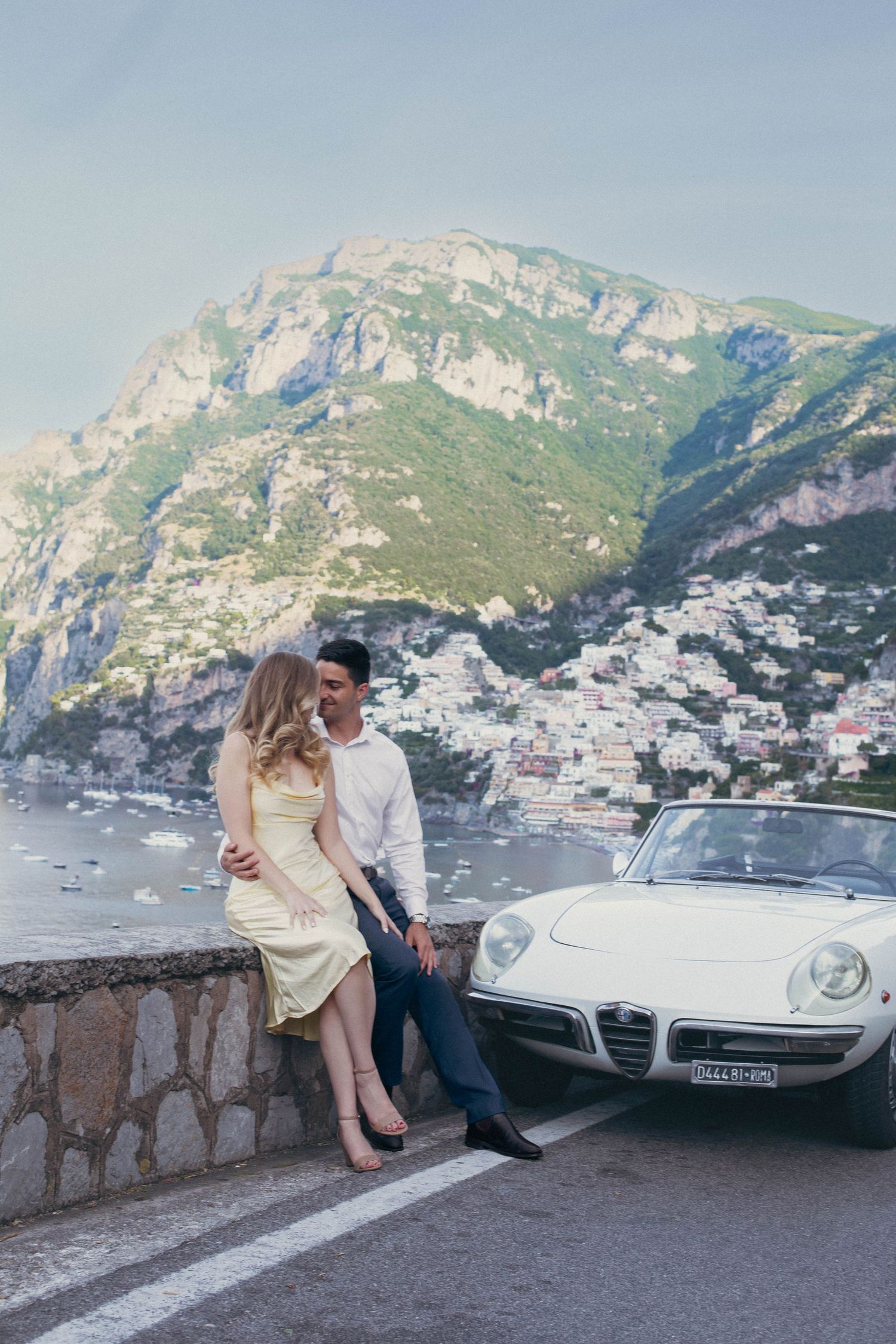 Couple poses by vintage Alfa Romeo convertible on Amalfi Coast road with Positano cliffside village backdrop.