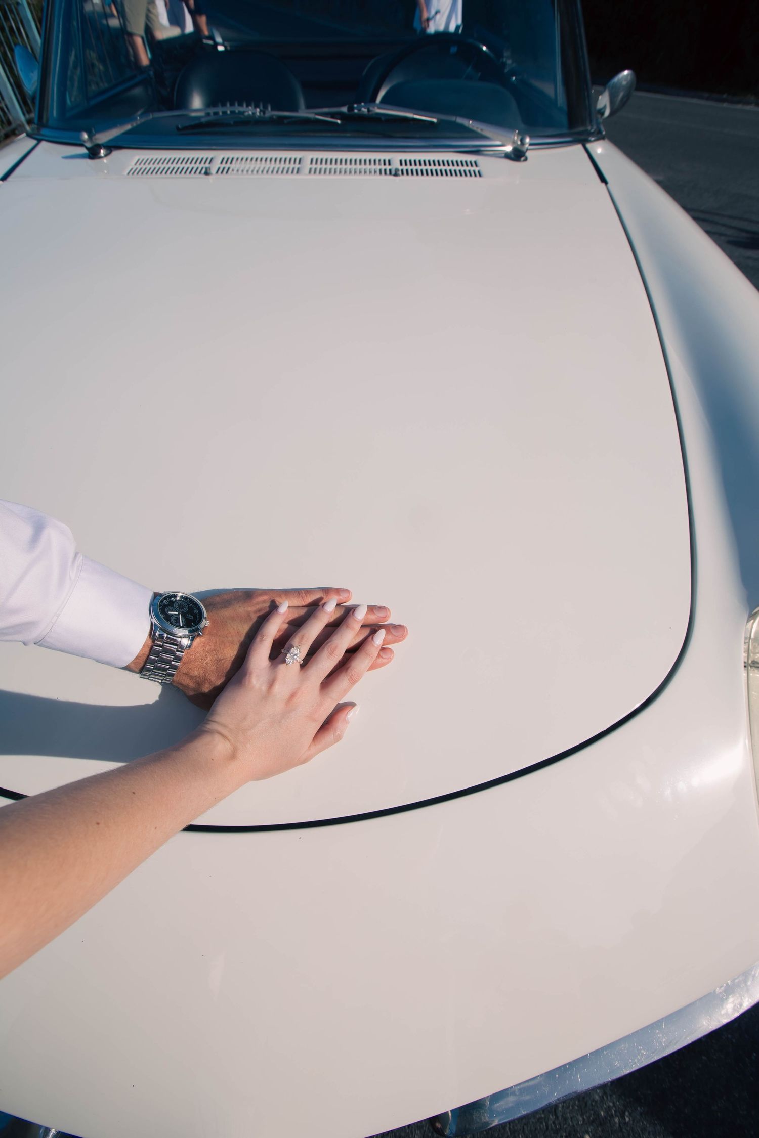 Two hands rest together on the hood of a classic white vintage car, one adorned with a silver watch and wedding ring.