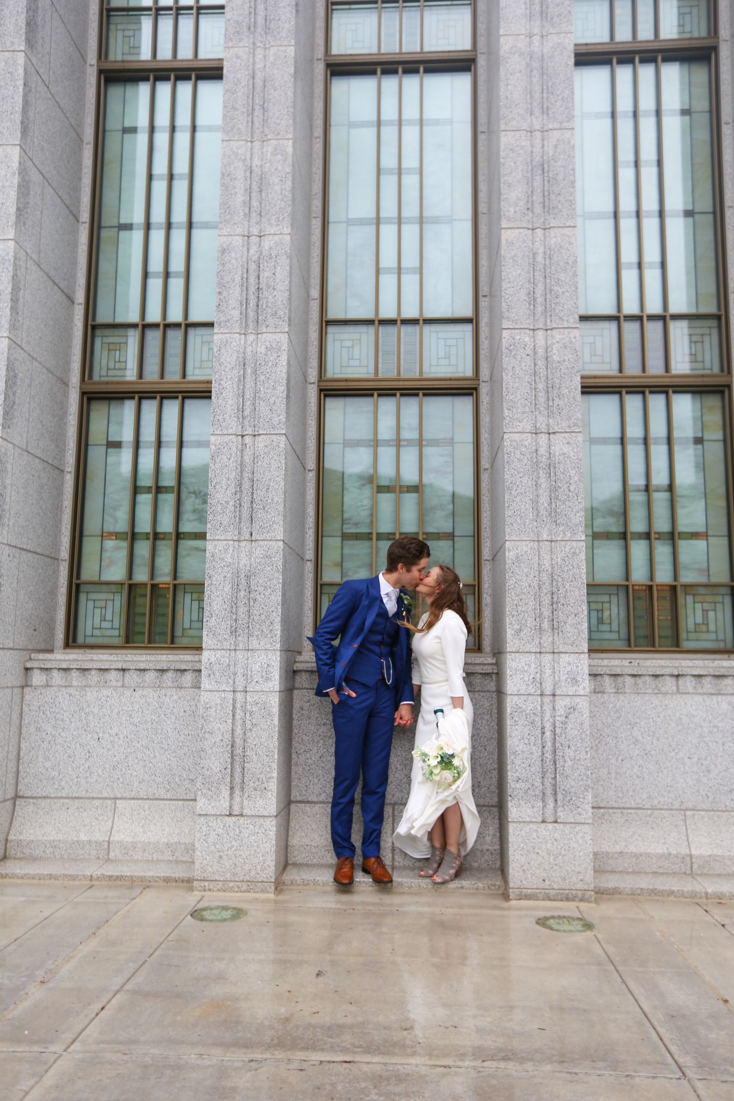 A couple poses against a modern stone building with tall windows for wedding portraits.