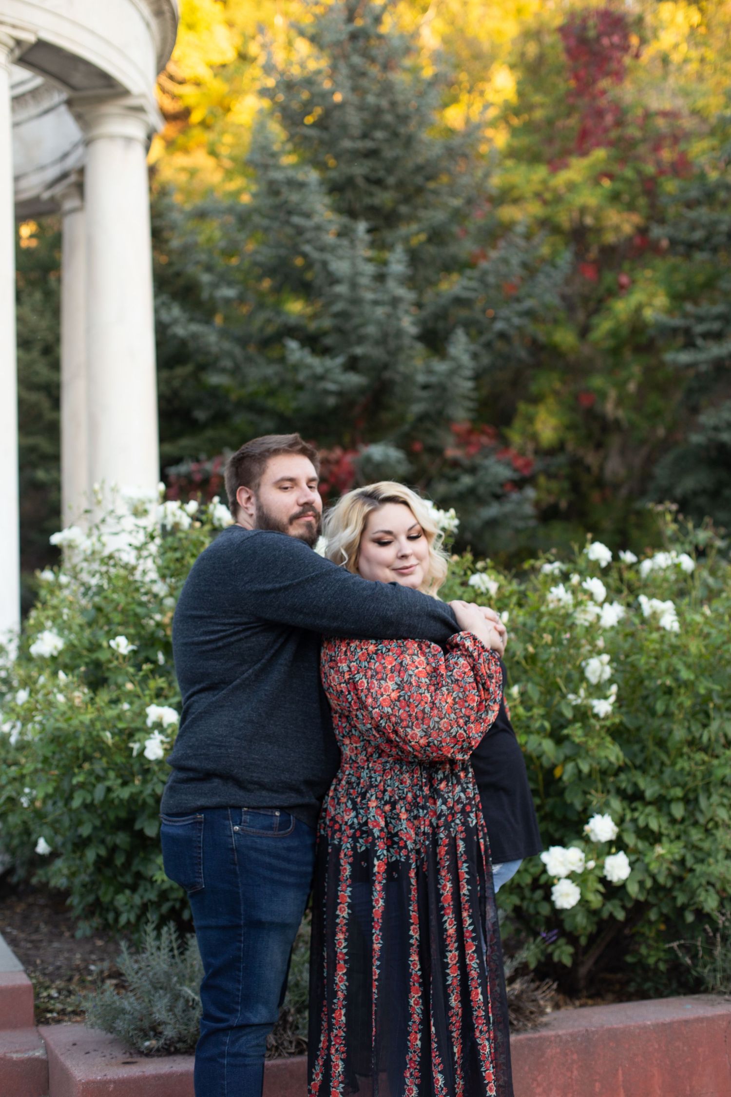 A couple embraces near white flowers and fall foliage by a garden gazebo.