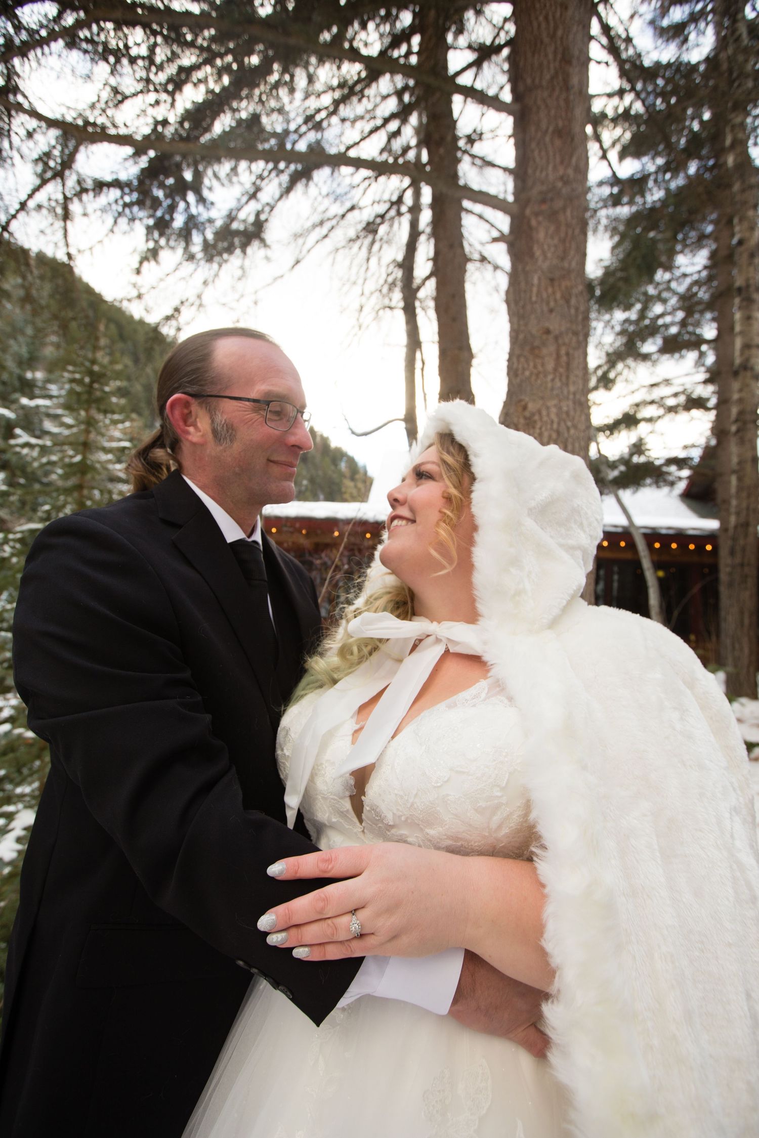 A joyful winter wedding portrait with snow-covered trees and a rustic cabin in the background.