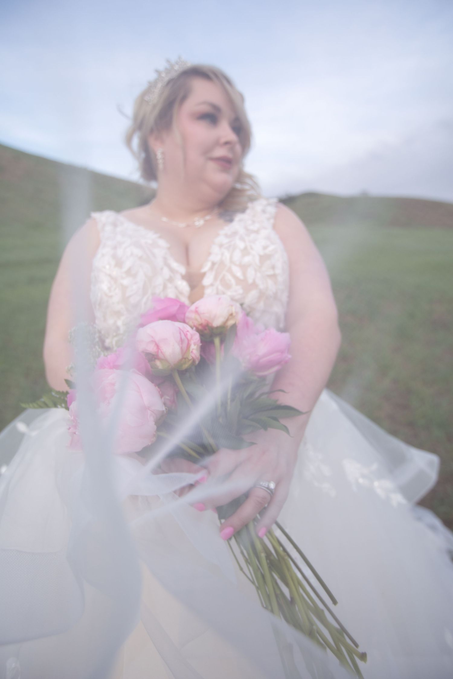 Ethereal bridal portrait with pink bouquet against soft blue sky and grassy hillside.