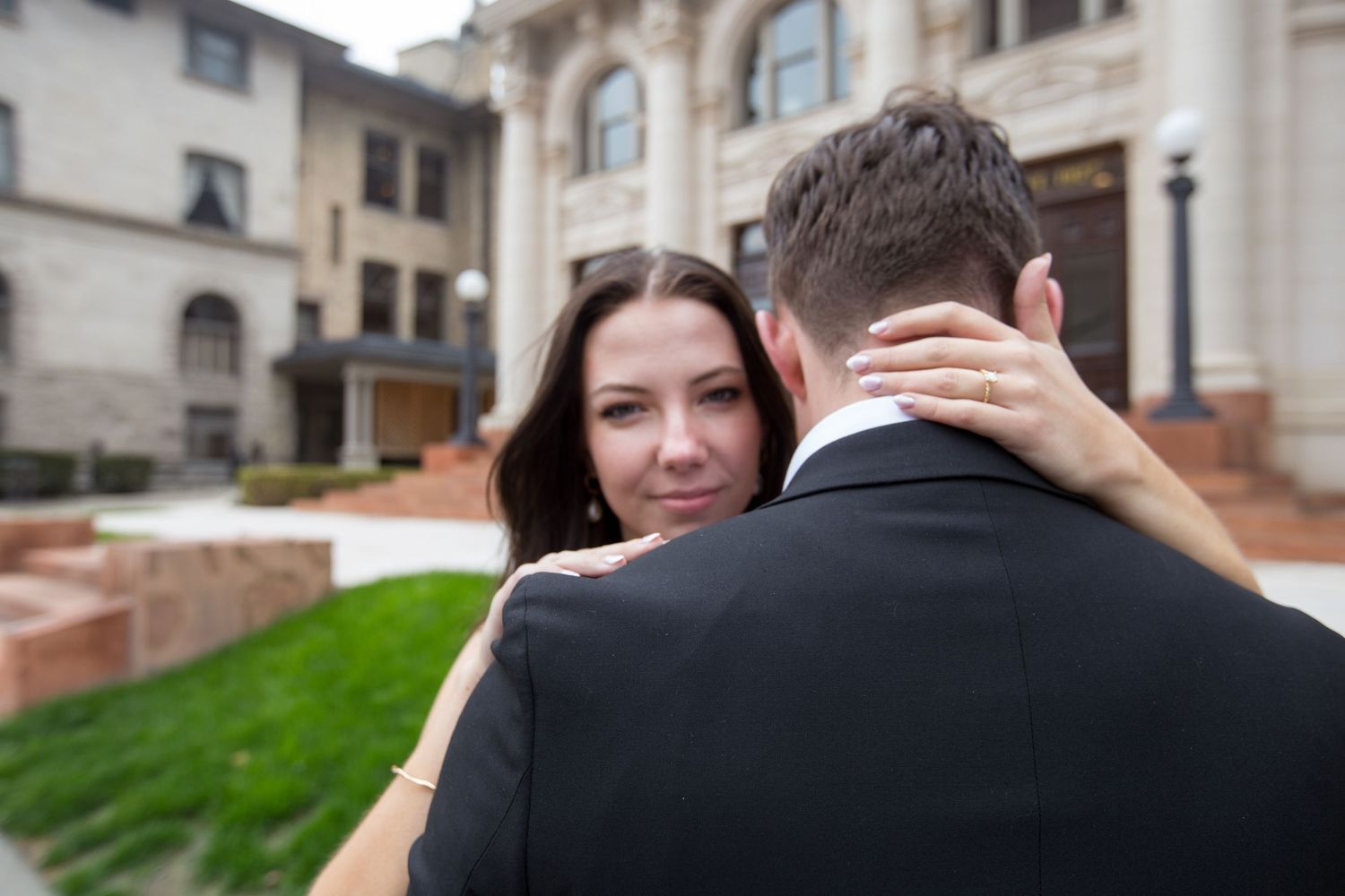 Bride embracing groom in front of elegant building, showcasing engagement ring during romantic outdoor wedding photo.
