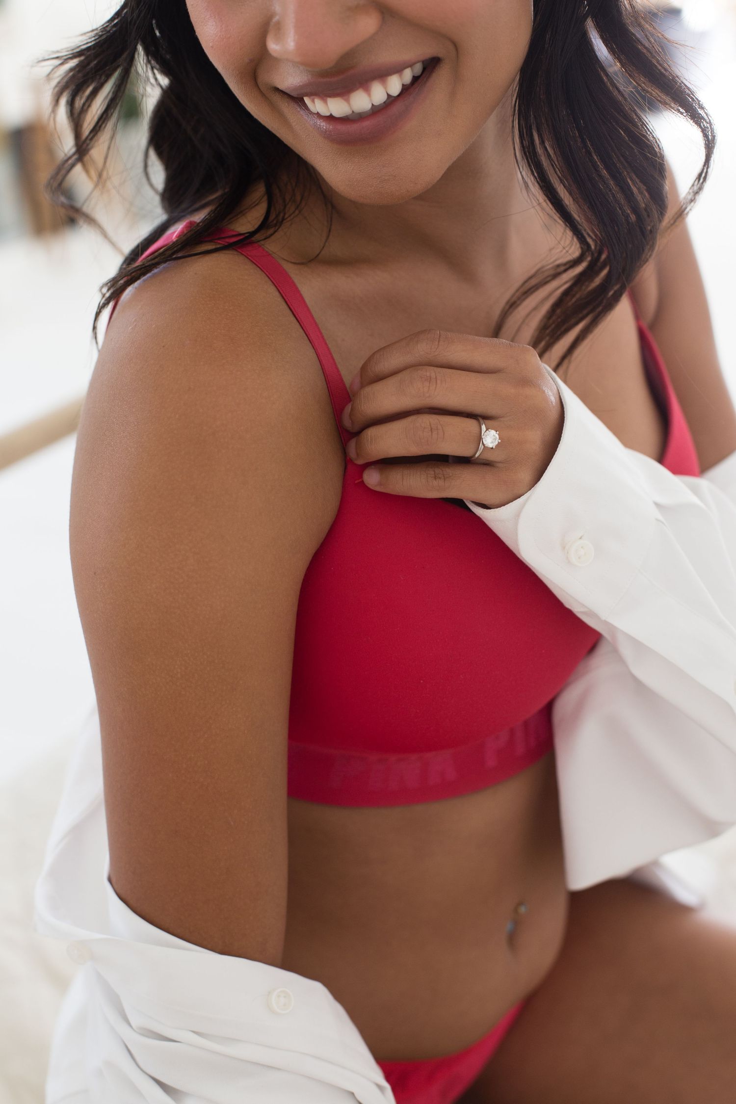 Smiling woman in red PINK lingerie bra wearing a diamond ring, partially draped in a white shirt.