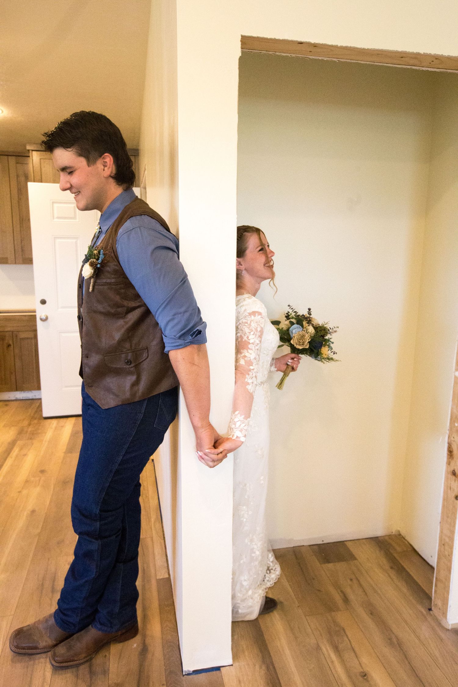 Groom in brown vest holds bride's hand around doorway as she smiles in white lace dress holding bouquet.