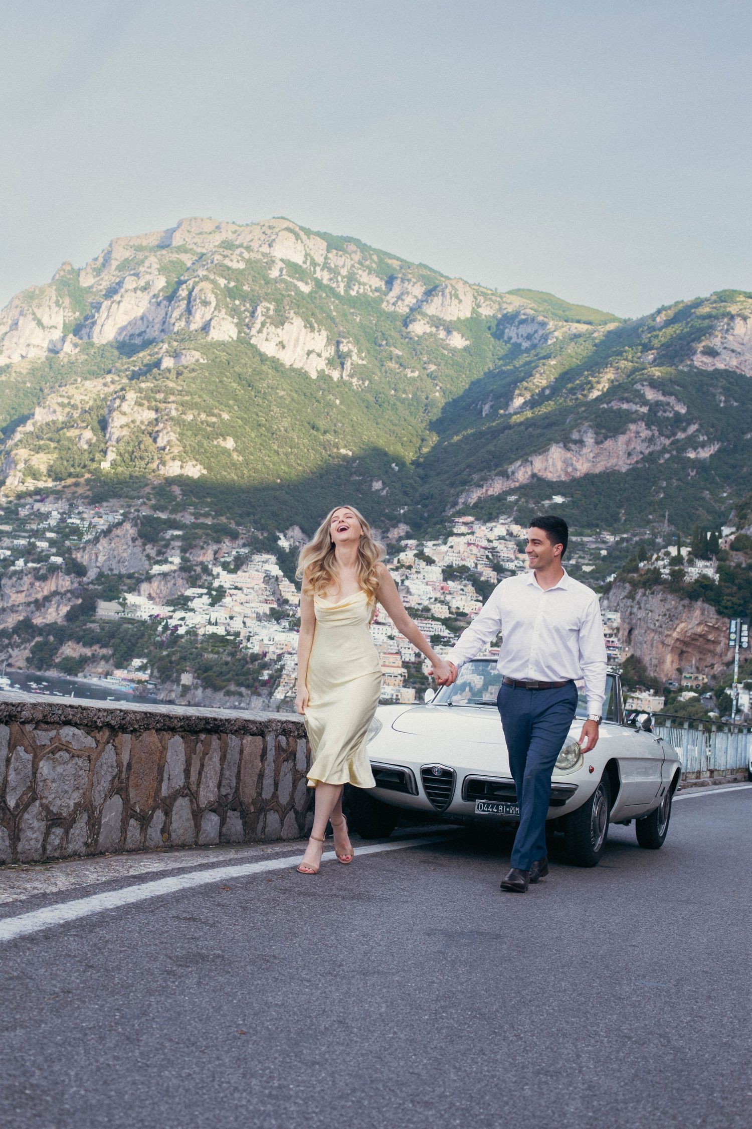 Elegant couple strolling hand in hand on scenic Amalfi Coast highway beside a classic white Alfa Romeo Spider.