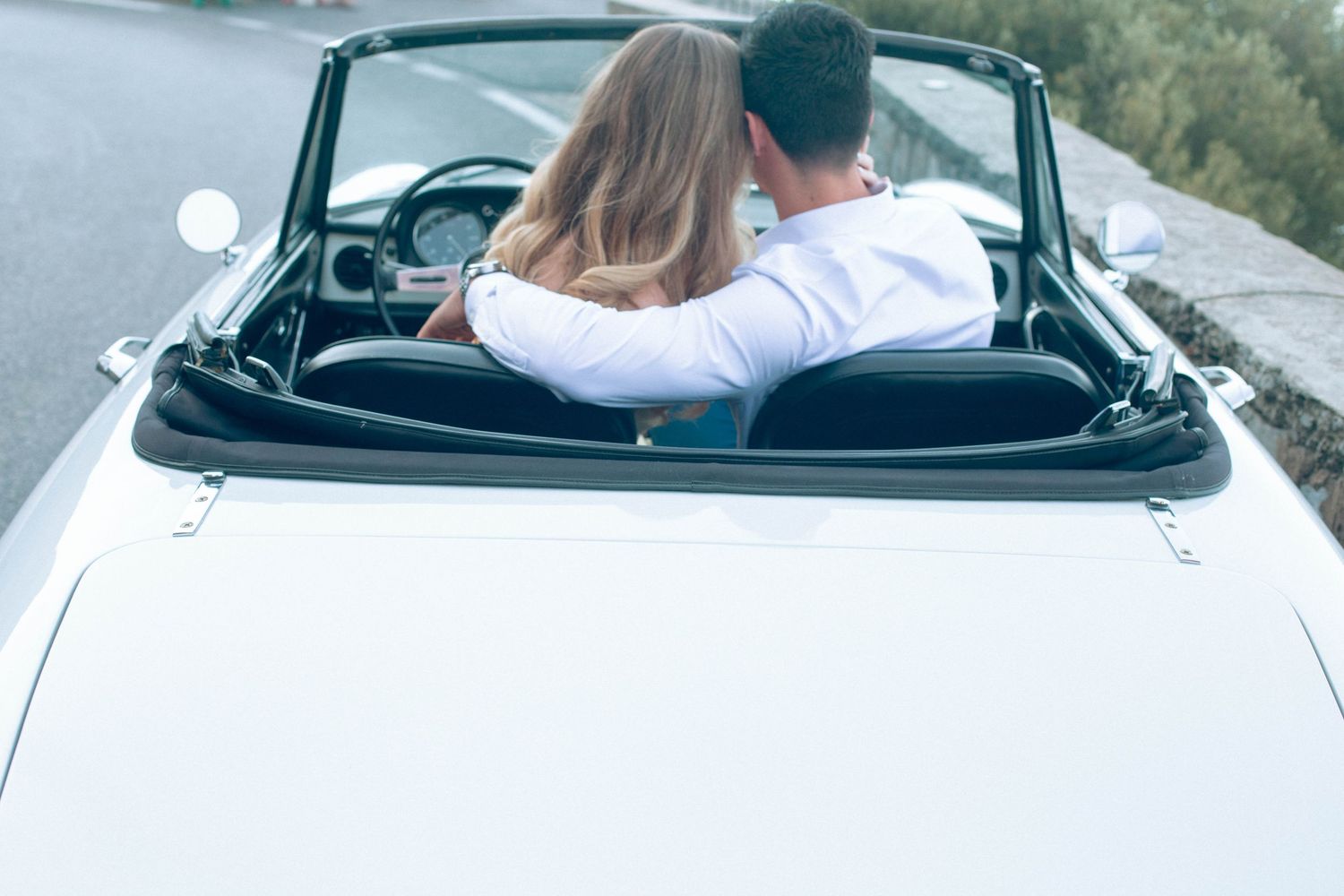 Couple embracing in white vintage convertible from behind on a scenic coastal road in Italy.