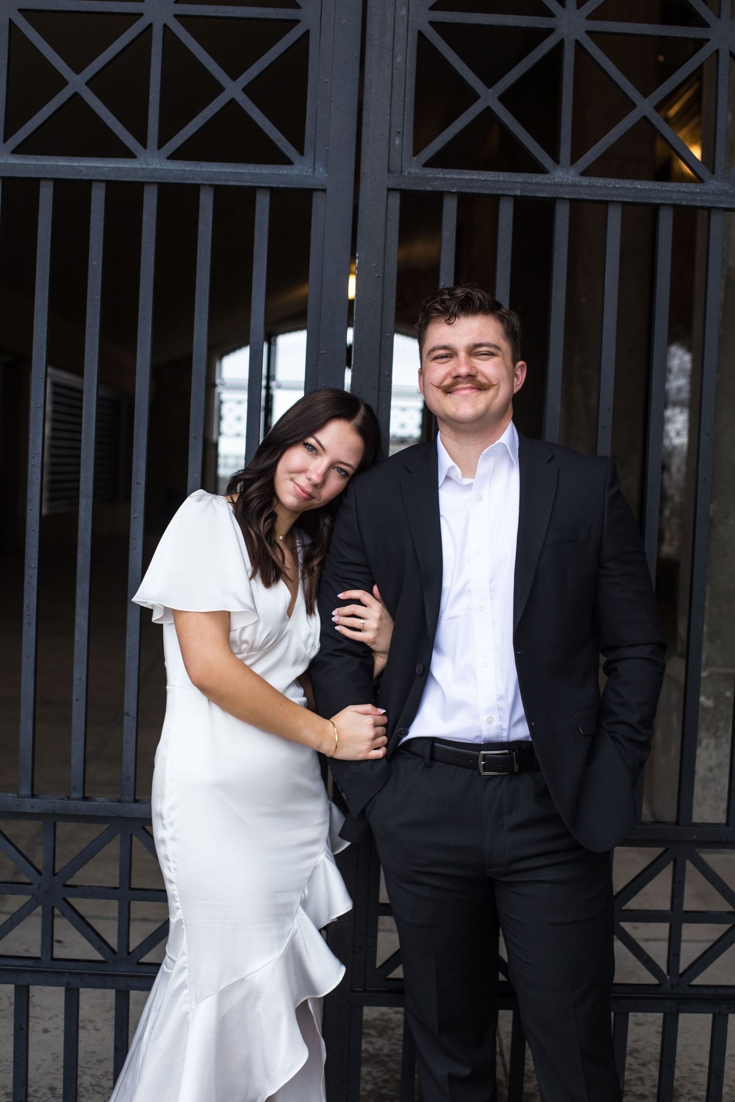A couple in formal attire poses playfully against an iron gate backdrop.