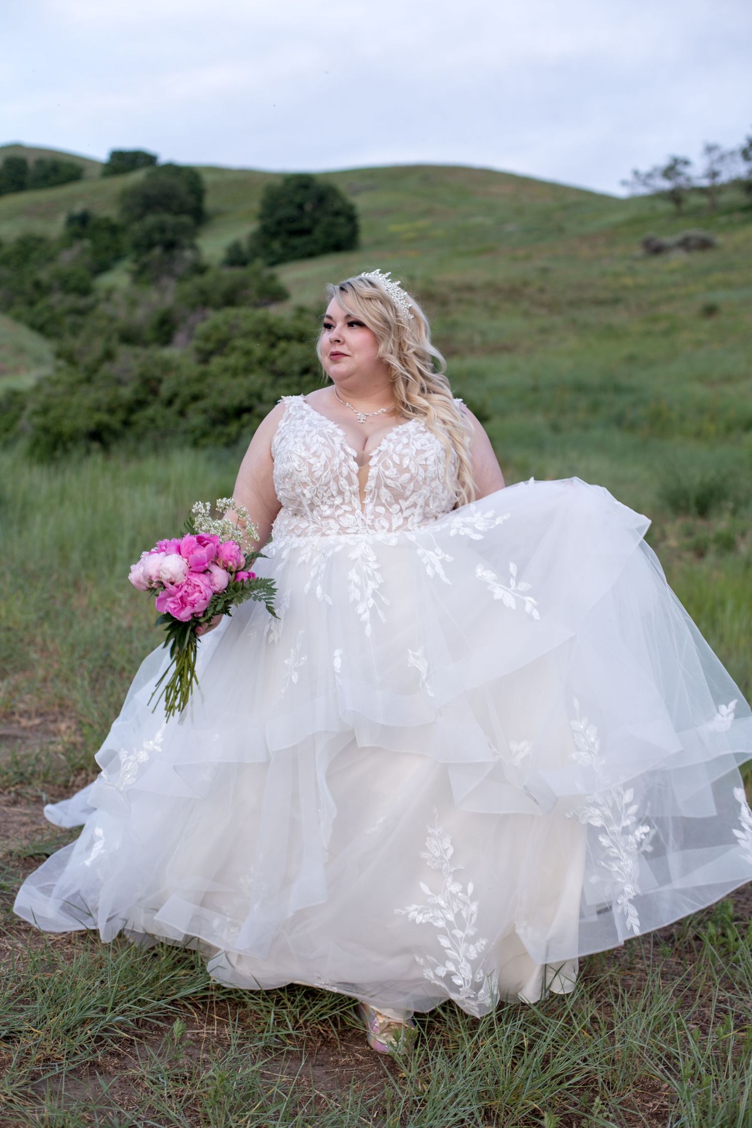 An elegant white wedding gown with lace details flows in the breeze on a grassy hillside.