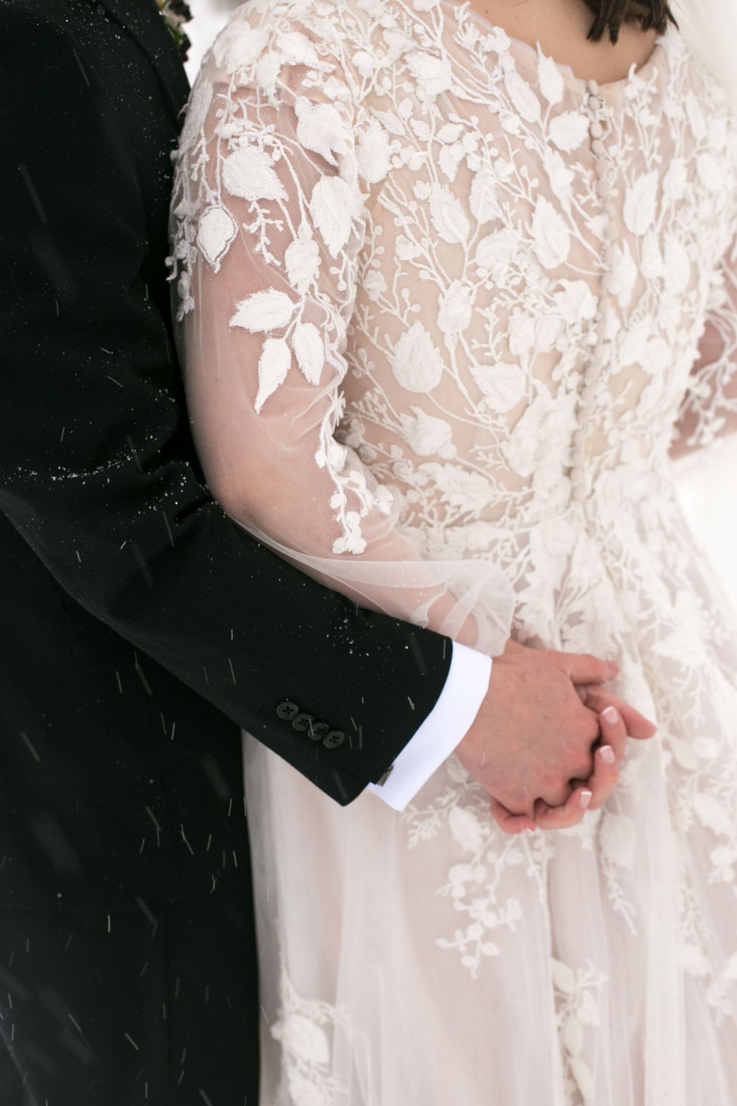 Close-up of delicate lace details on a white wedding dress sleeve with hands clasped together.