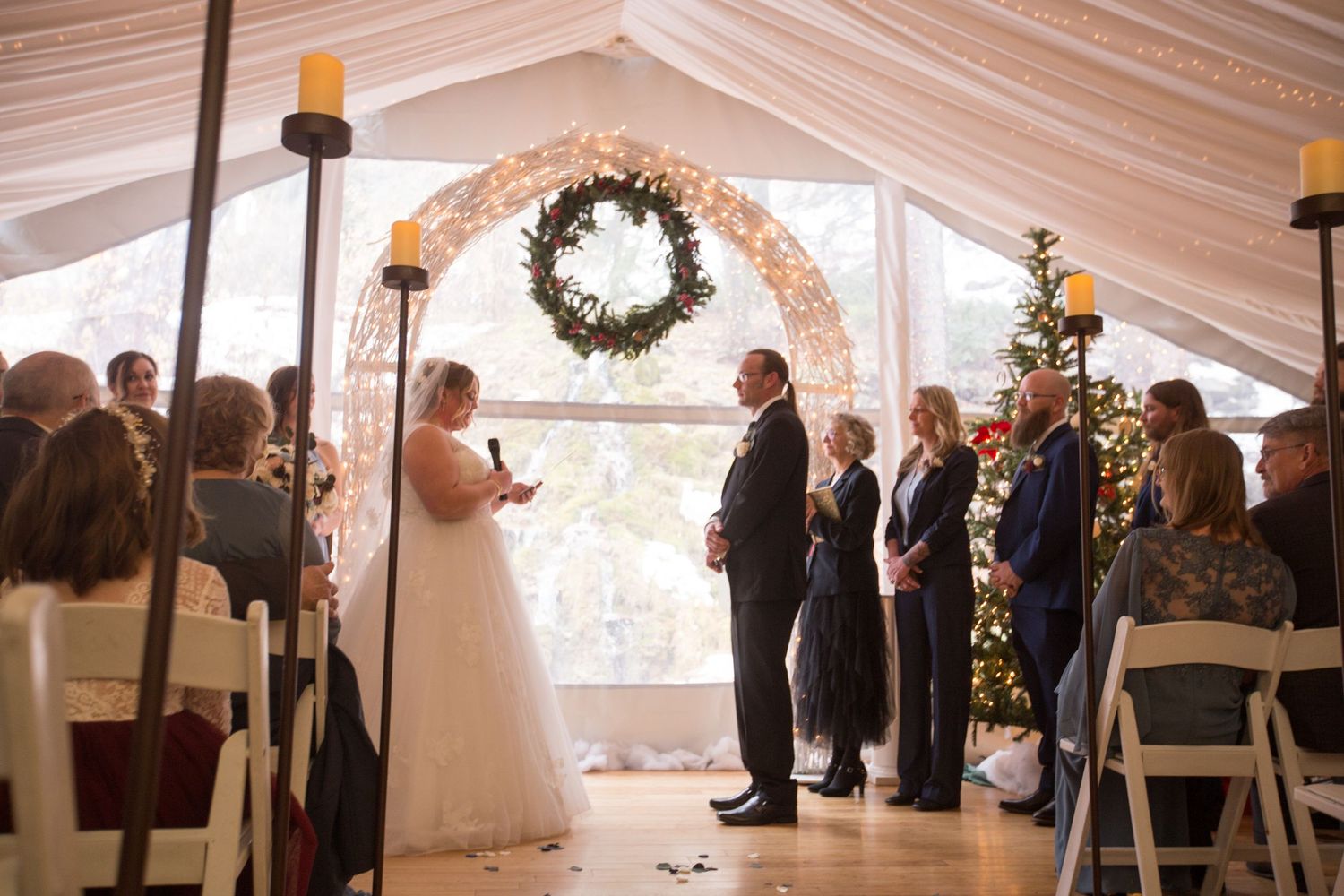 A bride and groom exchange vows at a Christmas-themed indoor wedding ceremony with a wreath arch and holiday decorations.