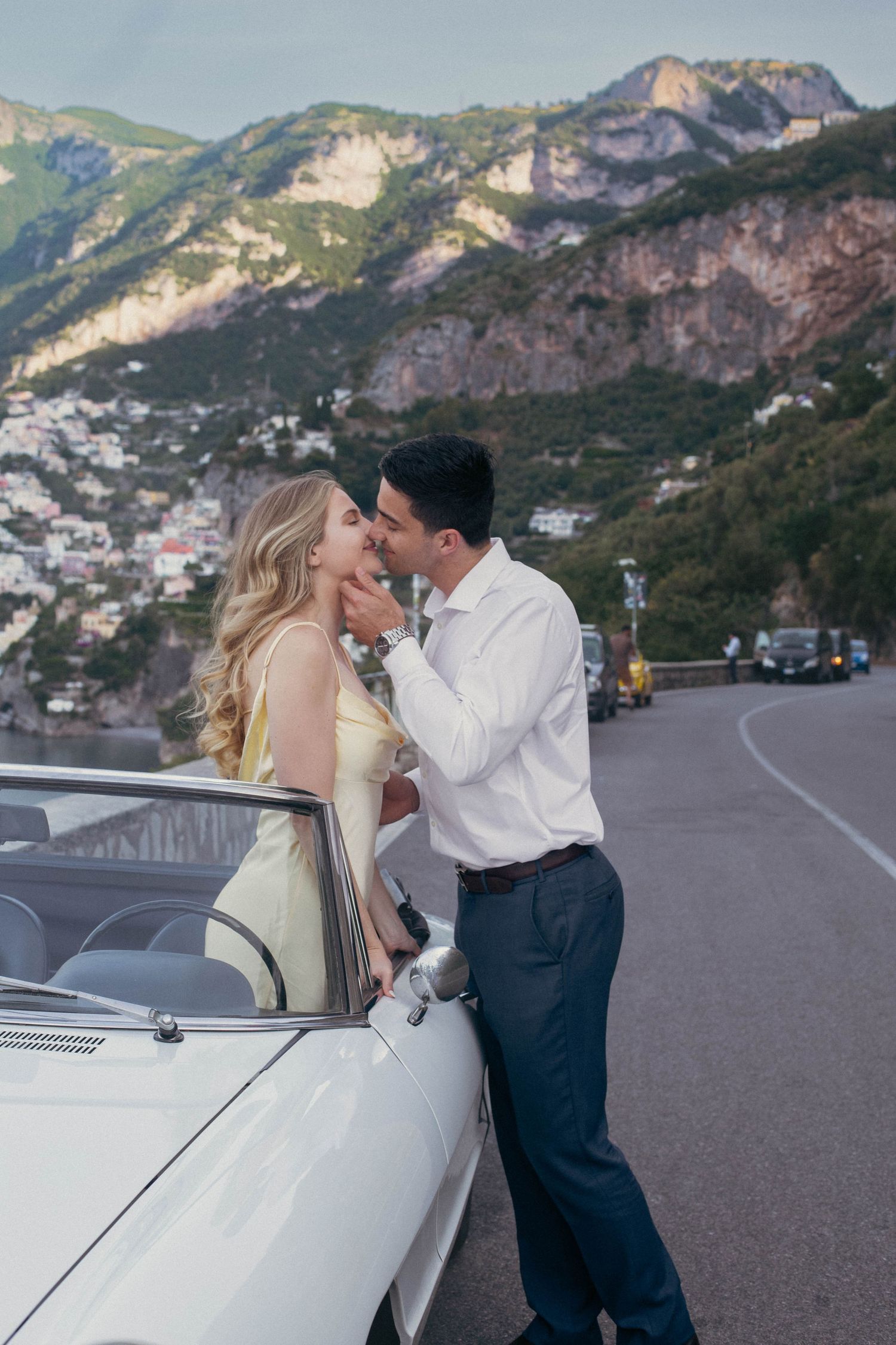 Couple sharing a kiss leaning against classic white convertible on scenic Amalfi Coast mountain road.