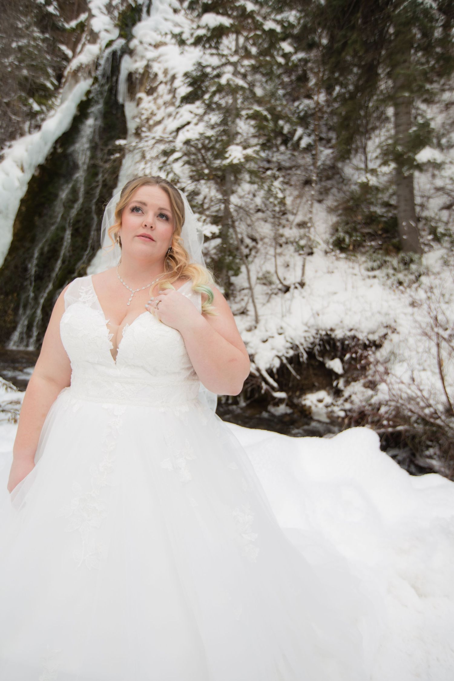 Bride in white ballgown stands gracefully against winter waterfall backdrop.