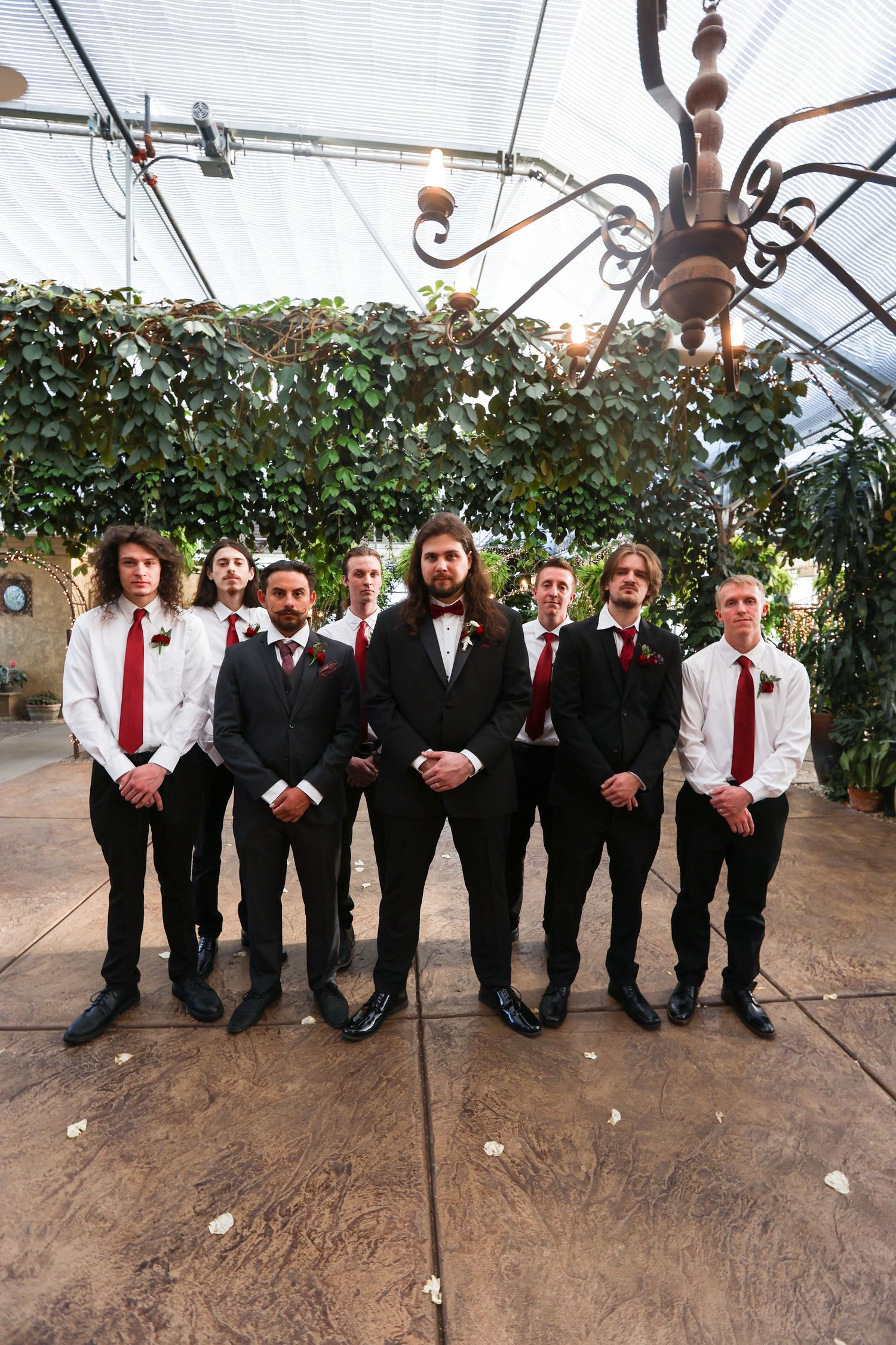 Groomsmen in black suits with red ties pose together in a greenhouse venue.