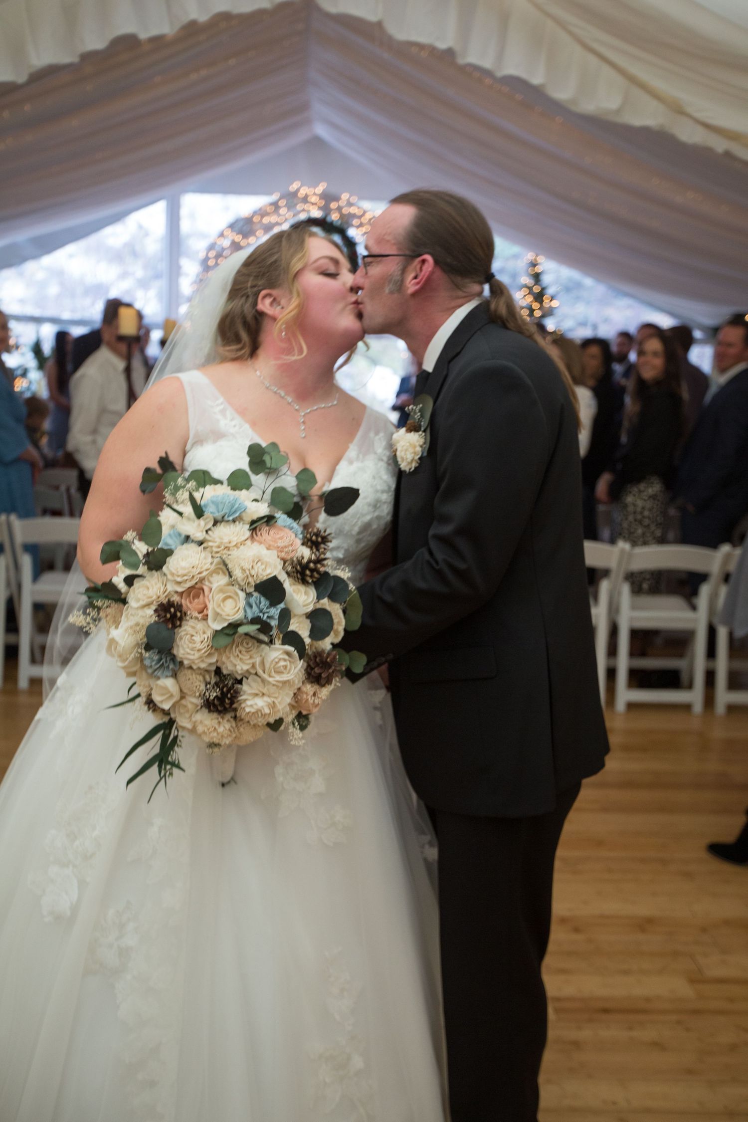 Bride and groom share first kiss at wedding ceremony in elegant white tent venue, bride holding rustic bouquet.
